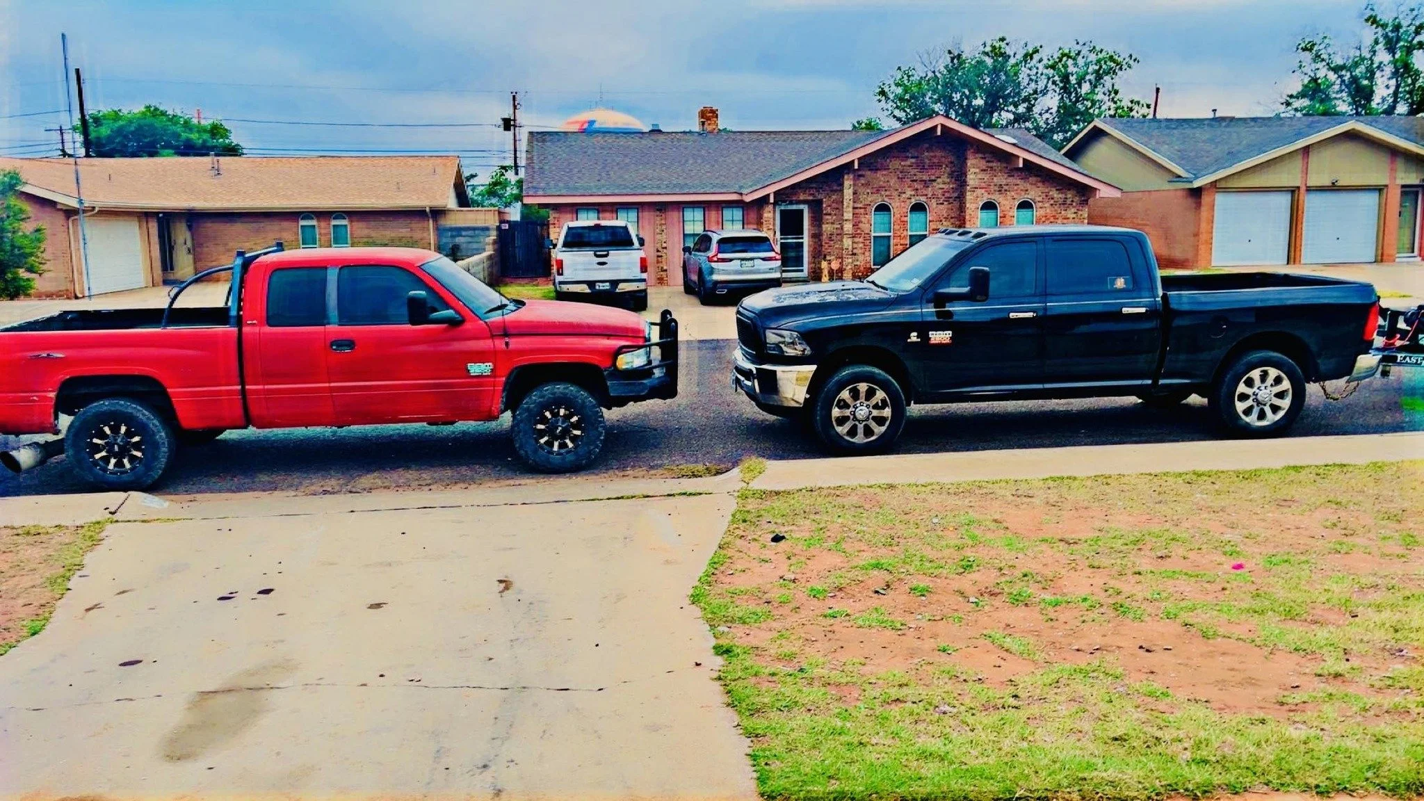 A suburban street with two pickup trucks parked in front of a row of single-family houses. The trucks are red and black, and the houses have brick exteriors and garages.