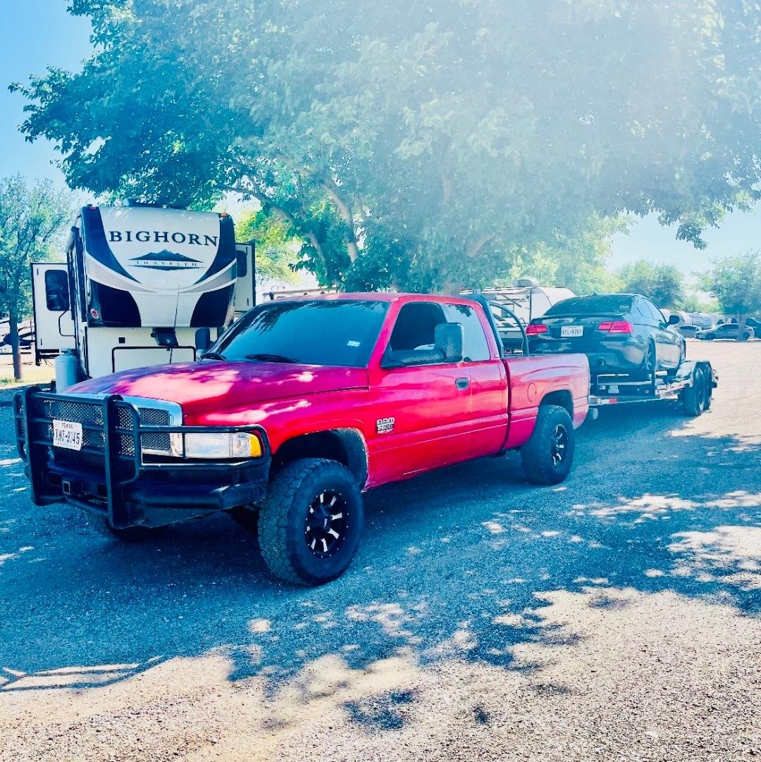 A red pickup truck towing a black car on a trailer, parked outdoors beneath a large tree with other vehicles and an RV in the background.