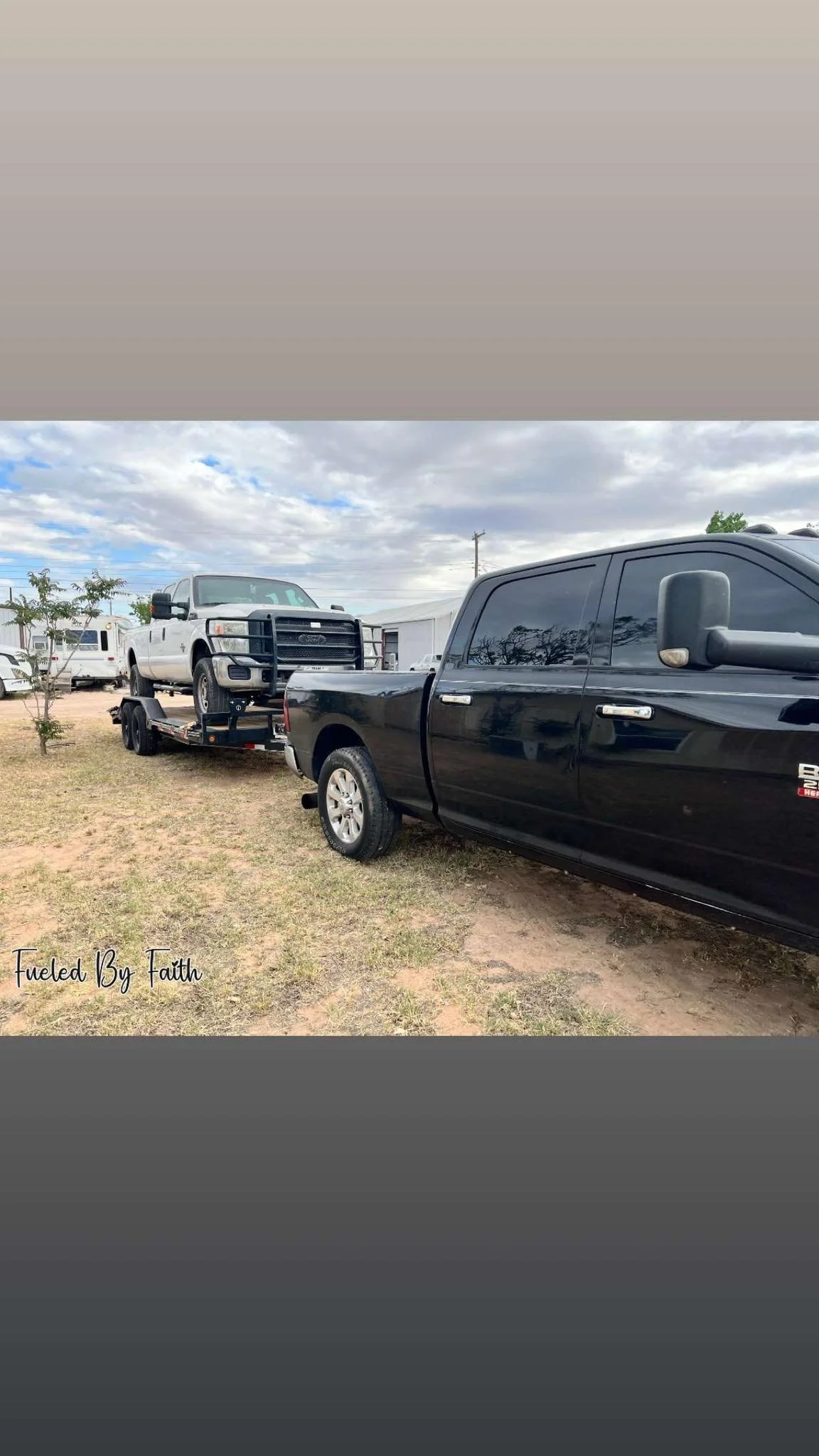 A black pickup truck towing a white pickup truck on a flatbed trailer in an outdoor lot with cloudy skies.