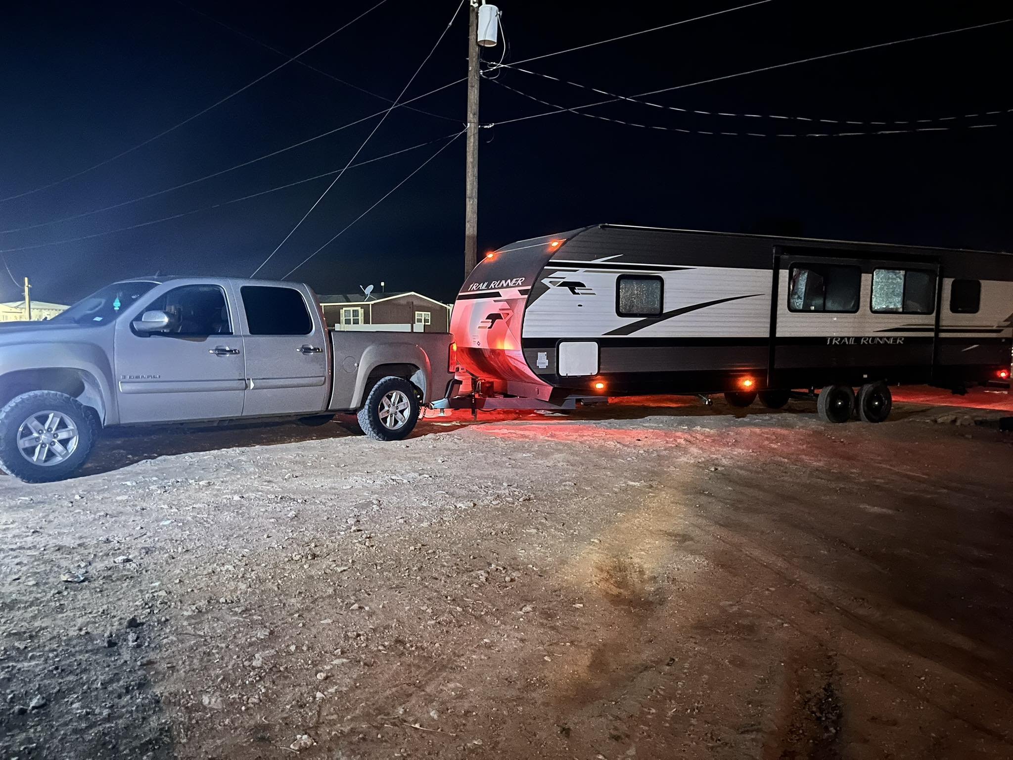 A pickup truck towing a travel trailer parked on a gravel surface at night, with overhead power lines and a house in the background.