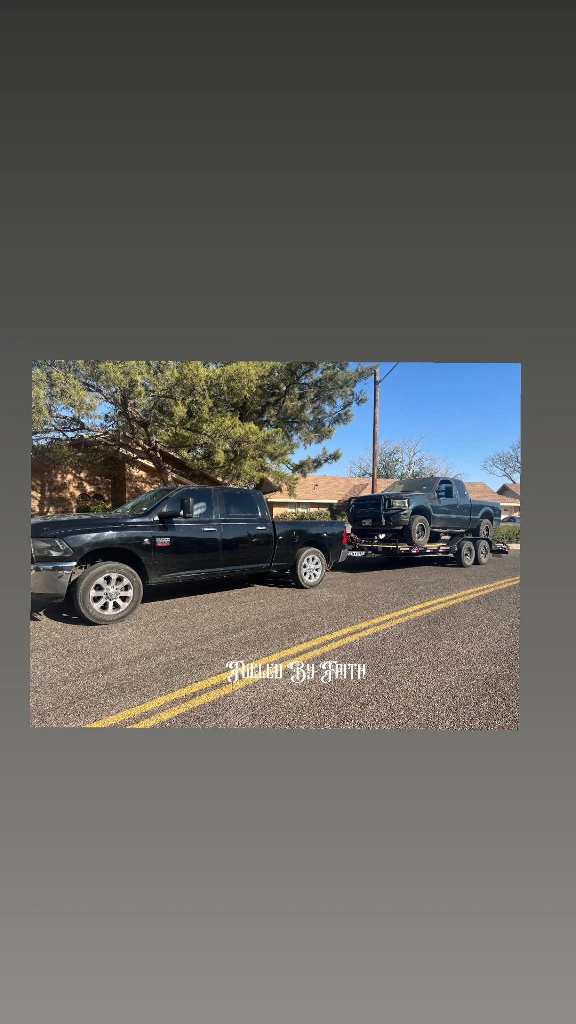 Black pickup truck towing a trailer with a black vehicle on it, parked on a street with houses and trees in the background.