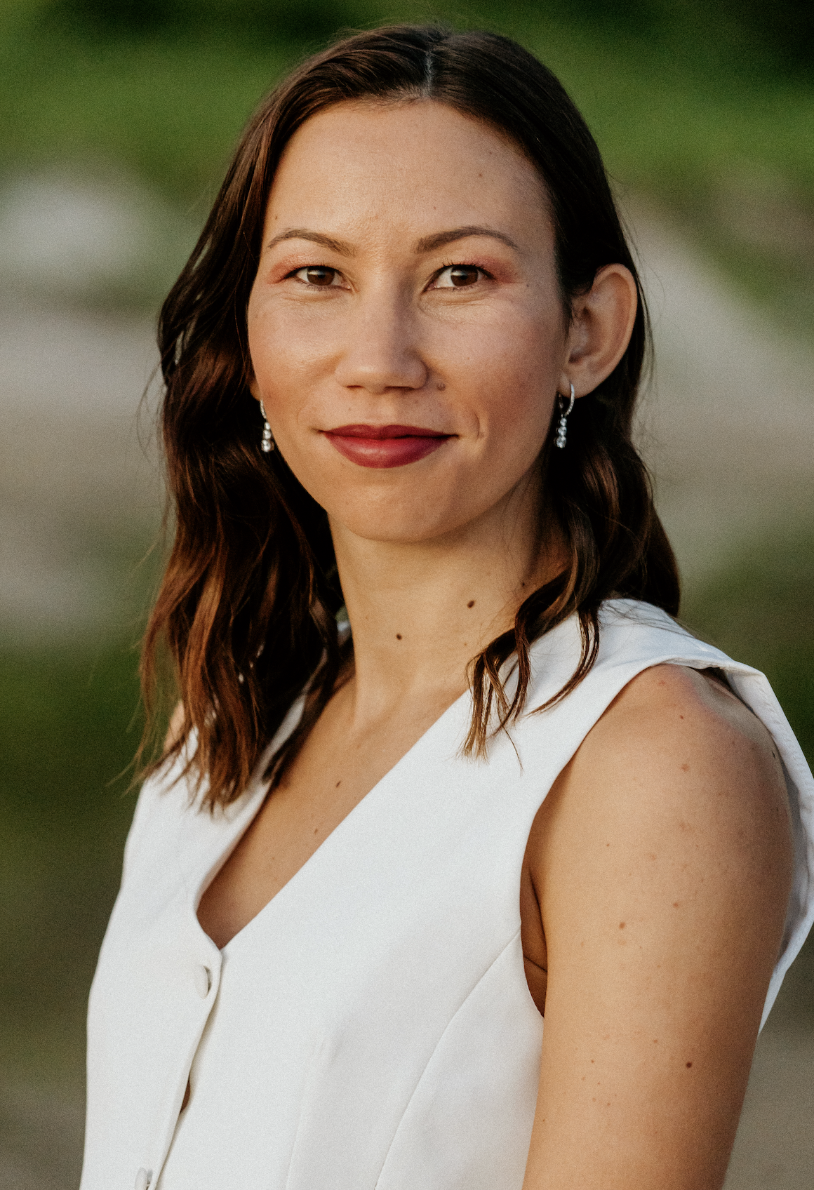 Portrait d'une femme souriante avec des cheveux bruns ondulés, portant des boucles d'oreilles et une robe blanche, en extérieur.