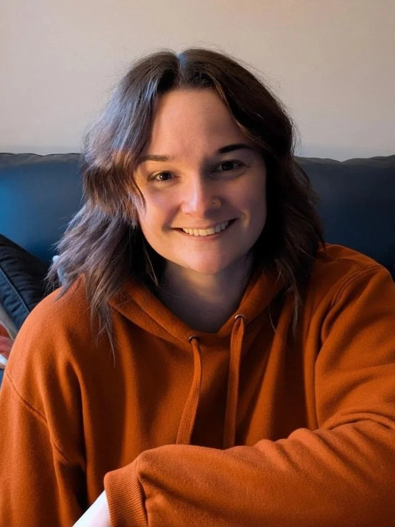 A young woman with shoulder-length brown hair smiling while sitting on a black couch, wearing a rust-colored hoodie.