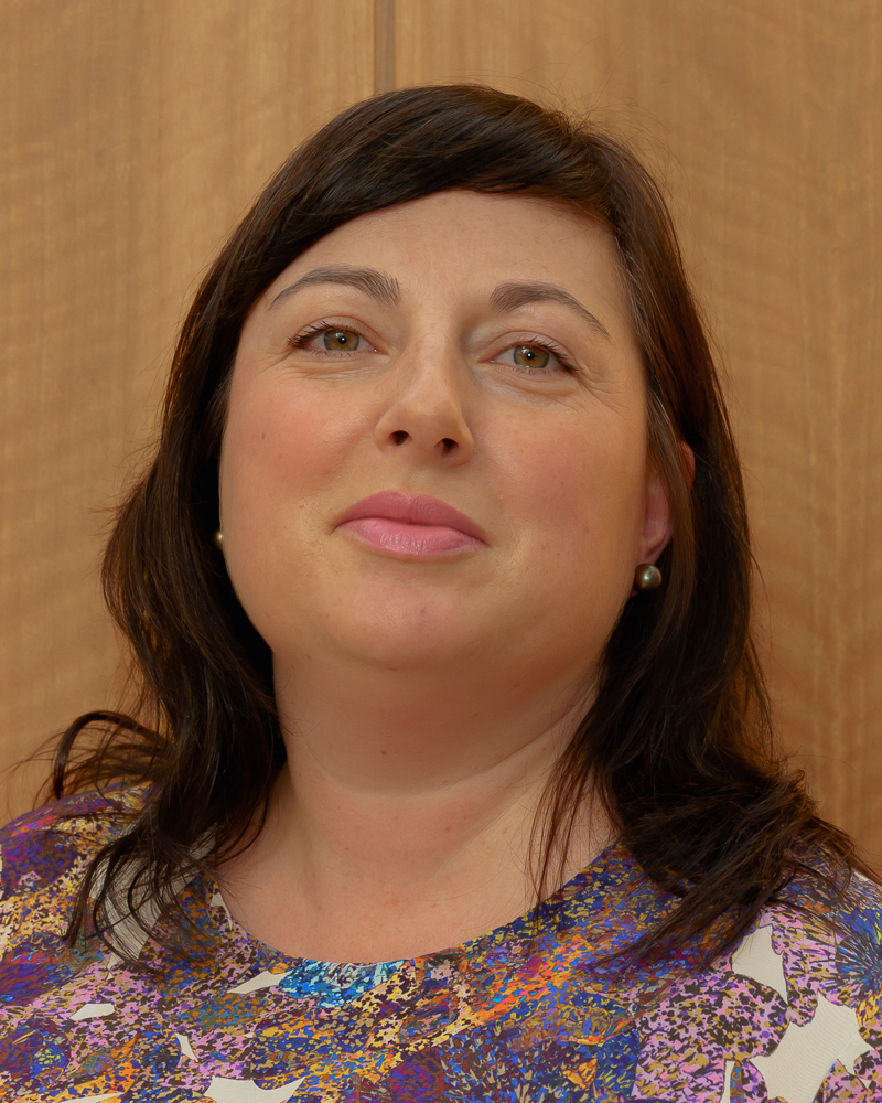 A woman with long brown hair smiles at the camera against a wooden background. This is Eliza Dawson, Advisor of Conservatory Residency and international arts residencies expert.