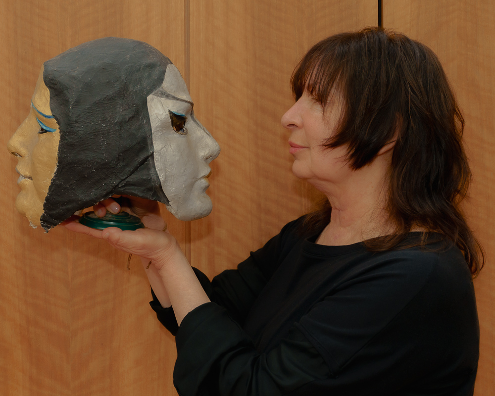 A woman with shoulder-length brown hair holds a papier-mâché mask of a woman's face, looking at it closely, against a wooden background. This is Léna Pangalo, Founder and Director of Conservatory Residency.