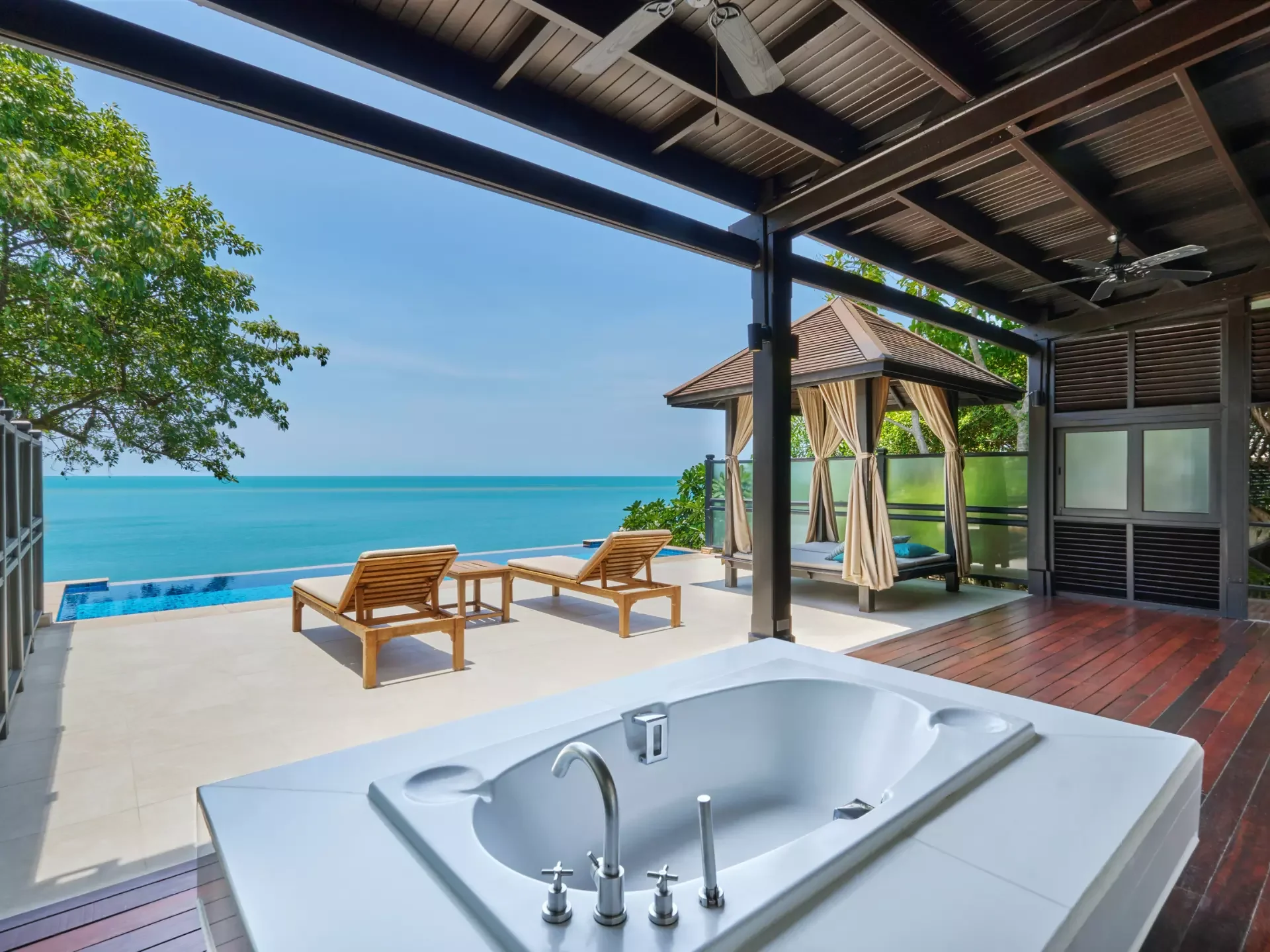 A view from a hot tub looking out onto a pool area with lounge chairs, and a cabana with curtains, overlooking the ocean with a clear blue sky.