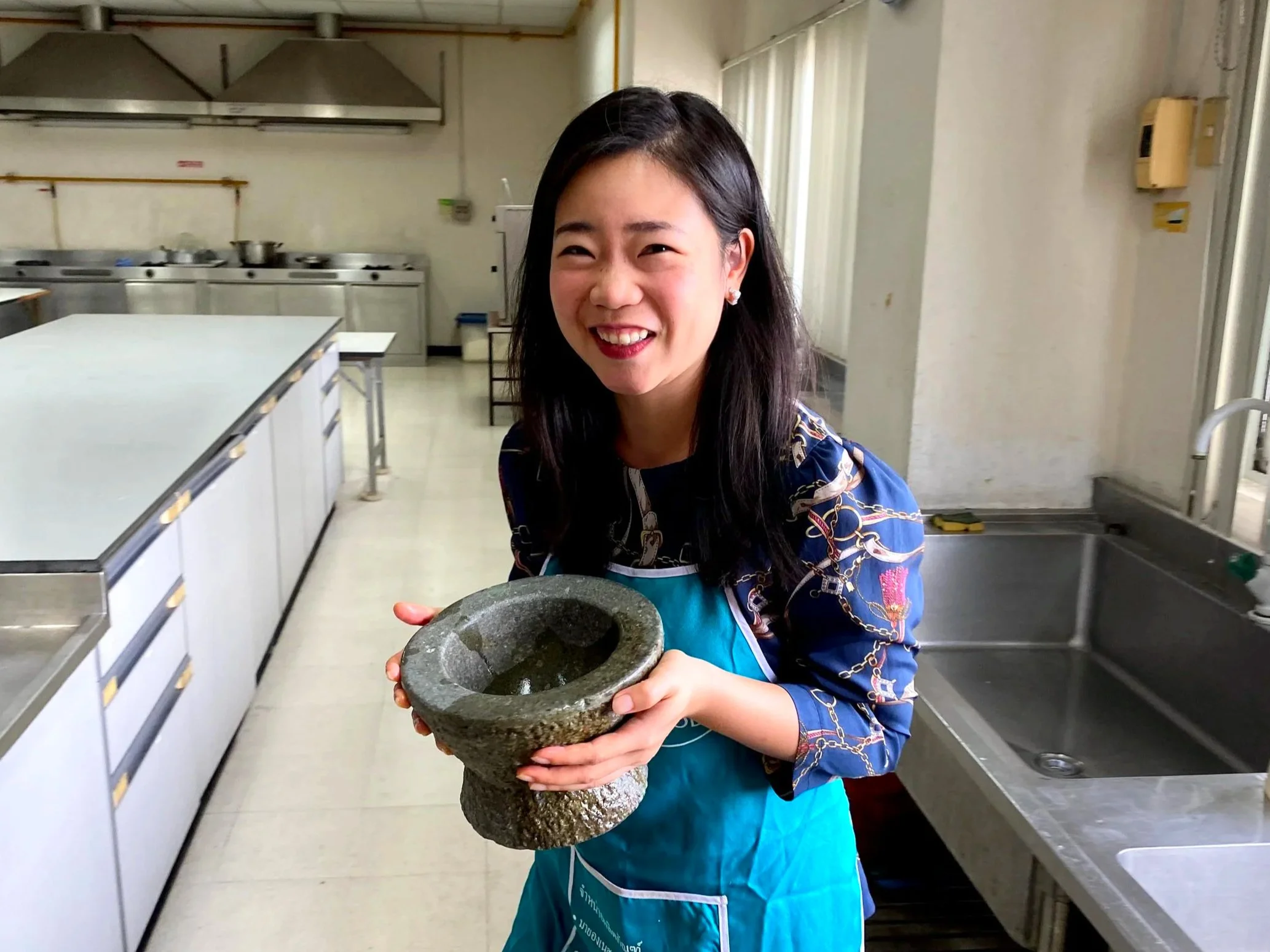 A woman with dark hair smiling and holding a black basalt grinder in a commercial kitchen.