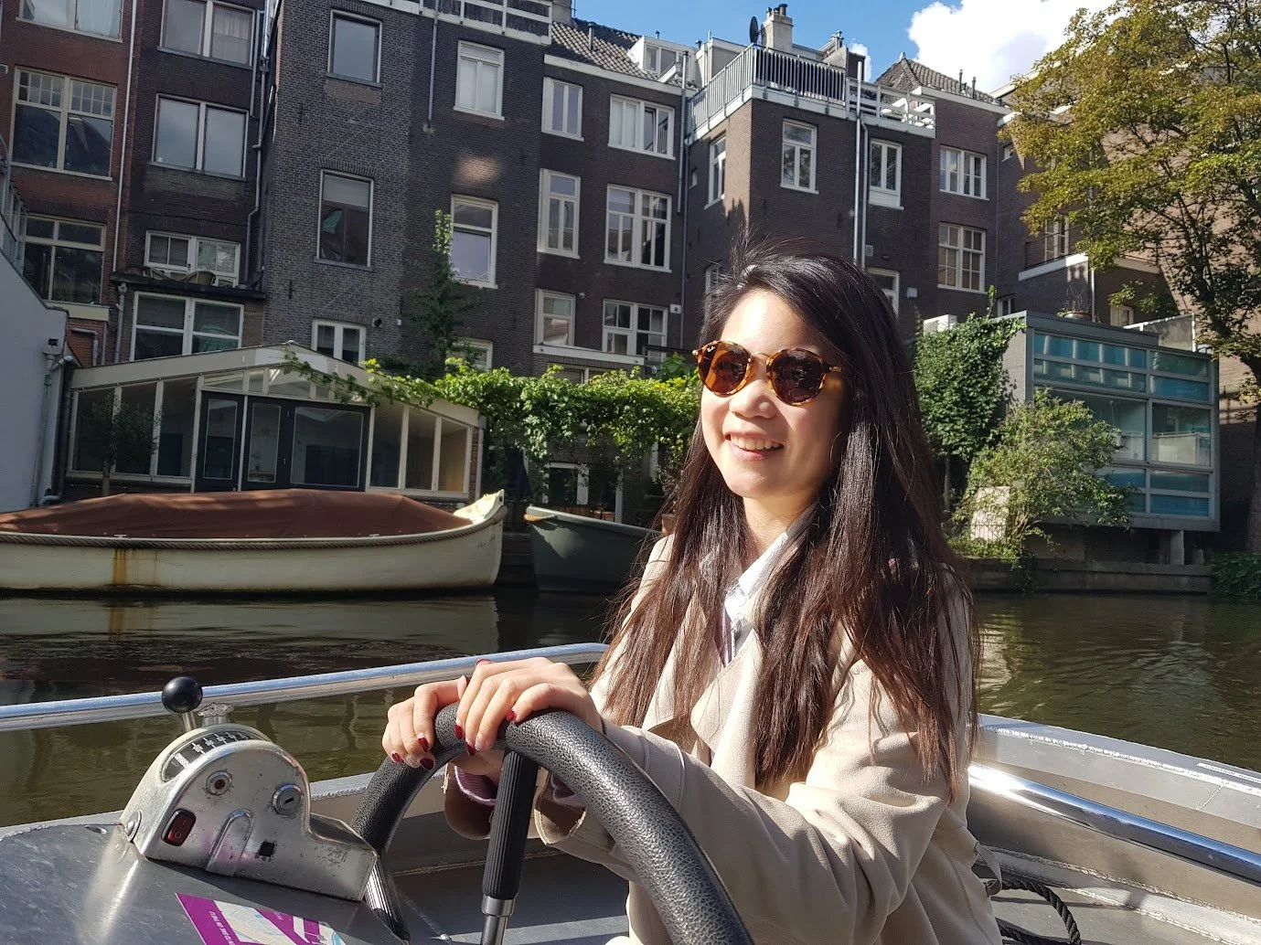 A woman with long dark hair and sunglasses smiling while steering a boat on a canal, with buildings and trees in the background.