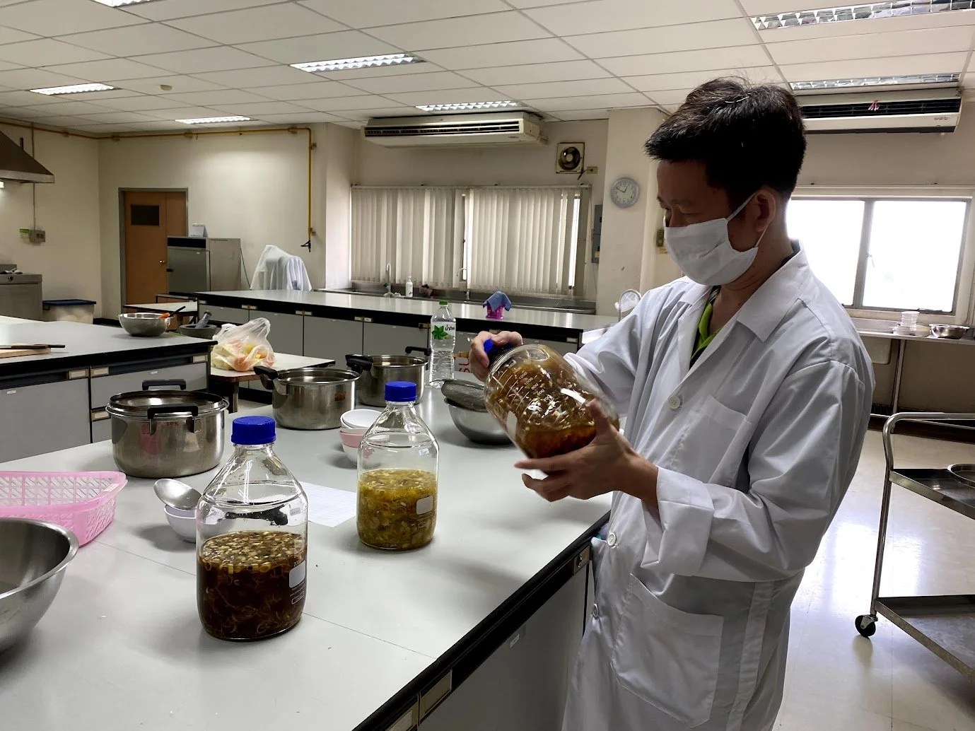 A man wearing a white lab coat and face mask is standing at a kitchen counter, holding a jar of fermented ingredients, with several jars of fermented items in front of him, in a clinical kitchen setting.