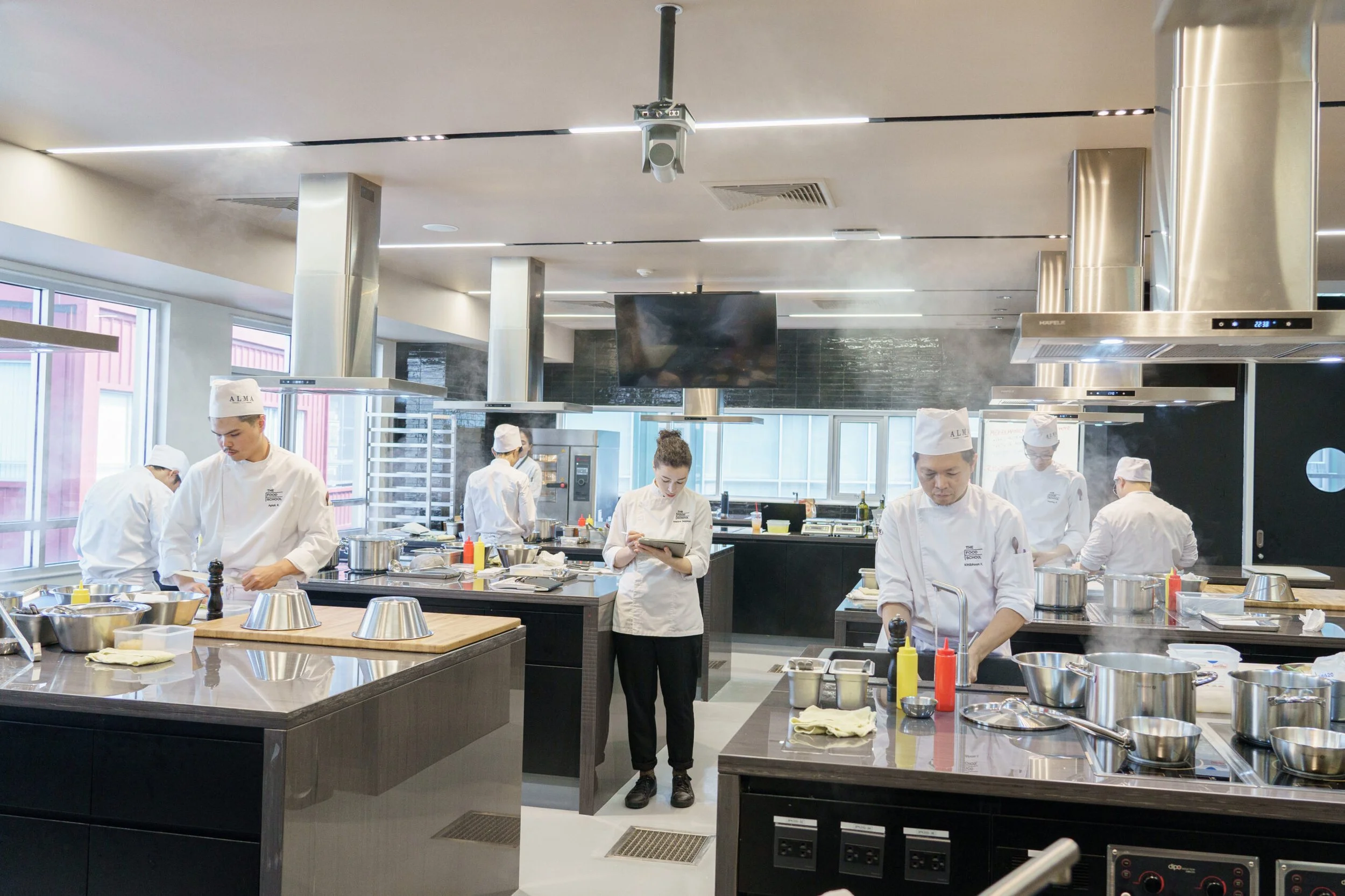 A professional culinary class taking place in a modern, well-lit kitchen with stainless steel appliances and large windows. Multiple students in chef uniforms are working at various stations, some reading recipes or preparing ingredients.