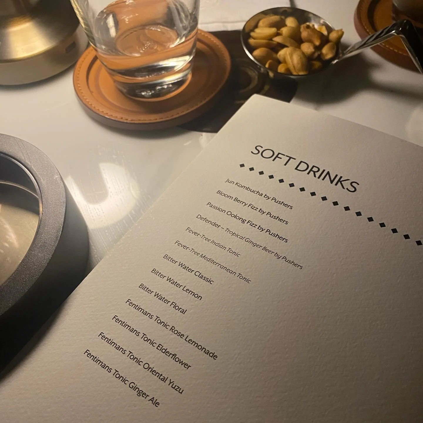 A menu titled 'Soft Drinks' on a table with a glass of water, a bowl of mixed nuts, and a metal pot. The menu lists various drinks including Jun Kombucha, Bloom Berry Fizz, Passion Oolong Fizz, Defender - Tropical Ginger Beer, Fever-Tree Indian Tonic, and others.