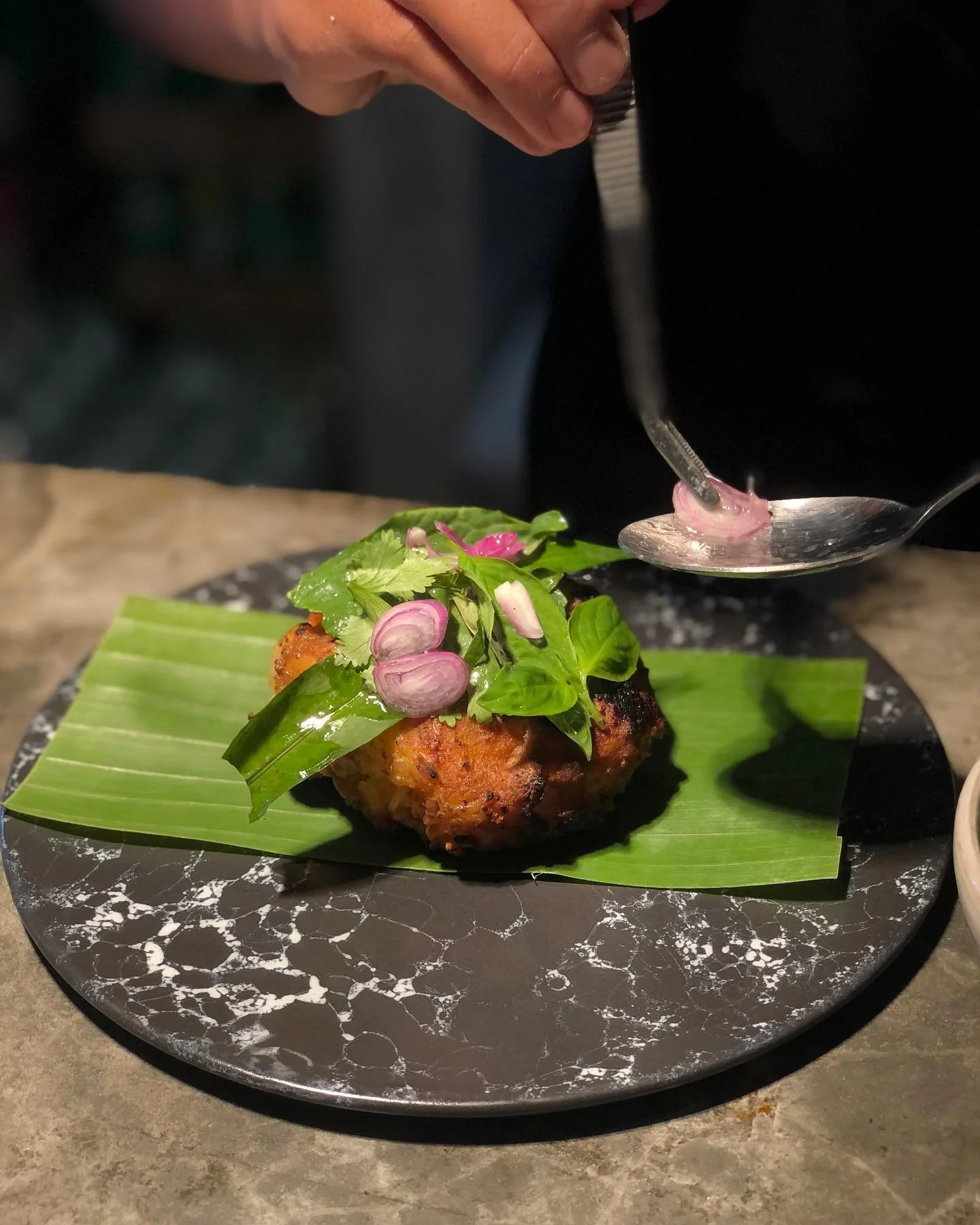 Fried dish topped with fresh herbs and sliced radishes on a black marble plate with a green banana leaf underneath, served on a gray stone table.