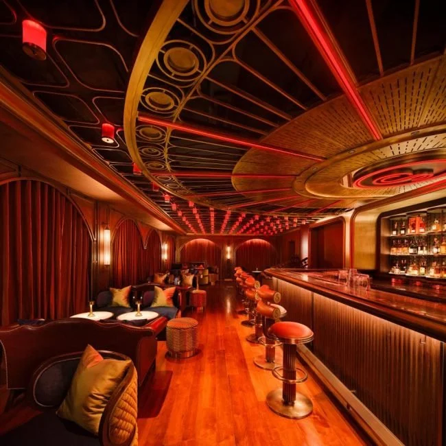 A cozy, dimly lit bar with a warm, inviting atmosphere, featuring a curved wooden ceiling with red neon accents, a long bar counter with high stools, and plush seating areas with cushions and small tables.