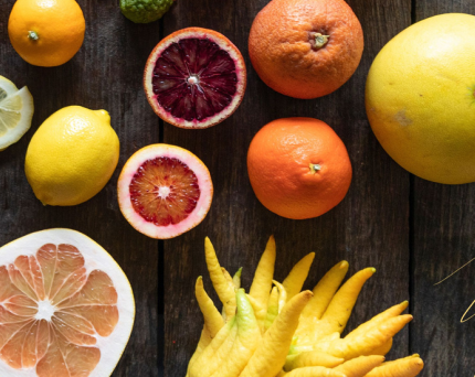 A variety of fresh citrus fruits on a dark wooden surface, including lemons, oranges, grapefruits, a blood orange, and yellow finger limes.