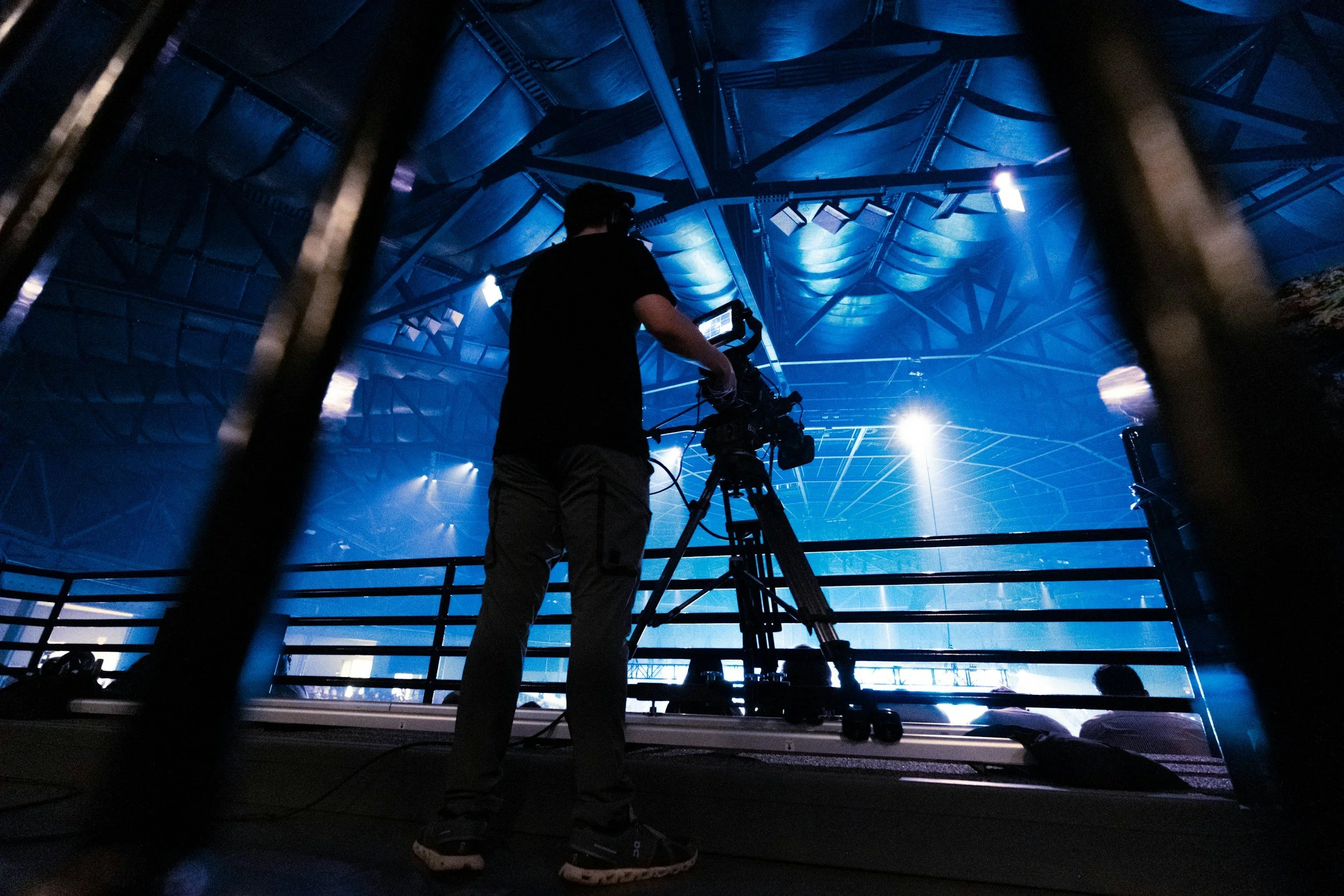 A person operating a camera on a tripod in a dark, industrial-looking room with blue lighting and a domed ceiling structure.