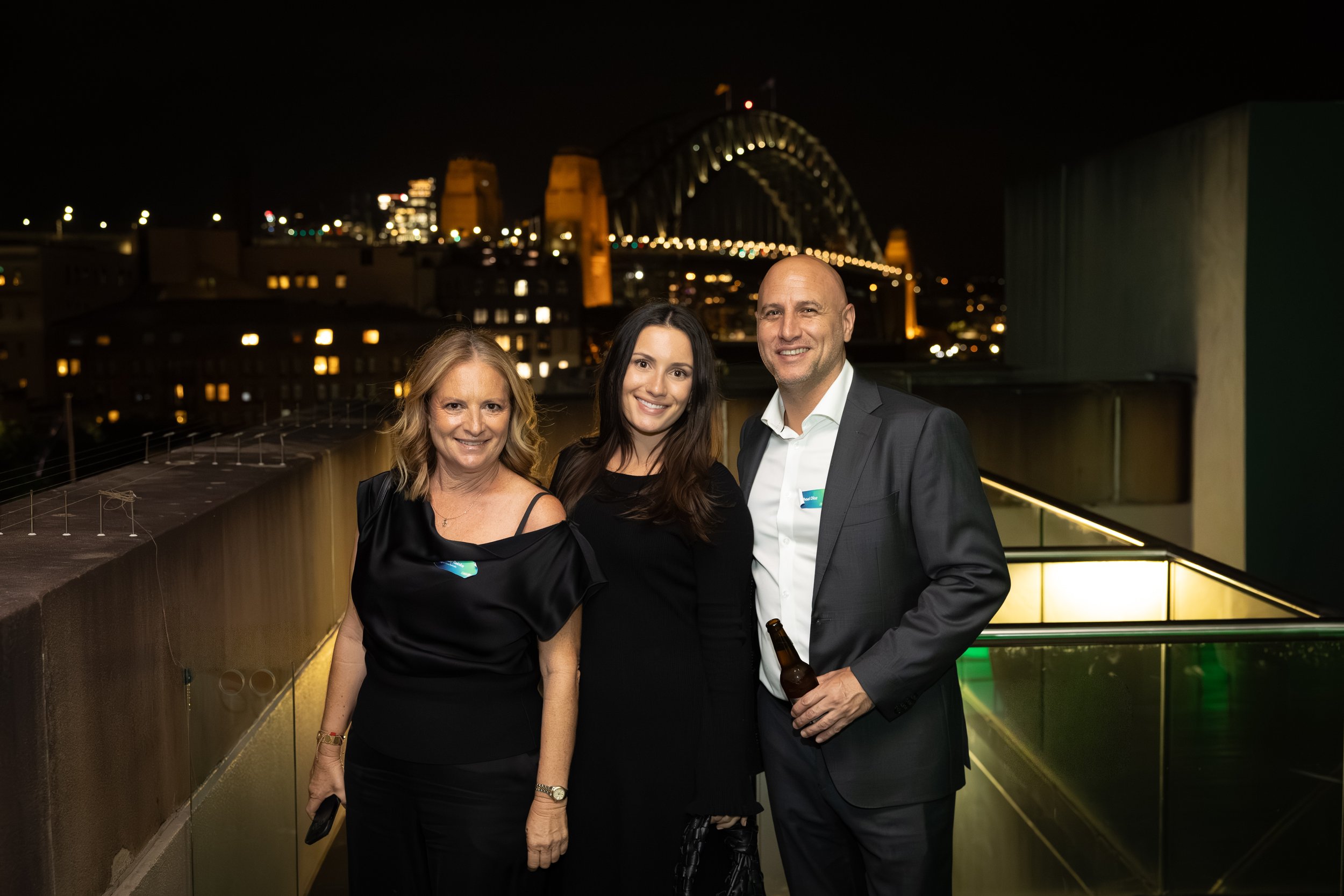 Three people standing on a balcony at night with city lights and a bridge in the background. Two women and one man, all dressed in formal attire, smiling at the camera. The man is holding a beer bottle.