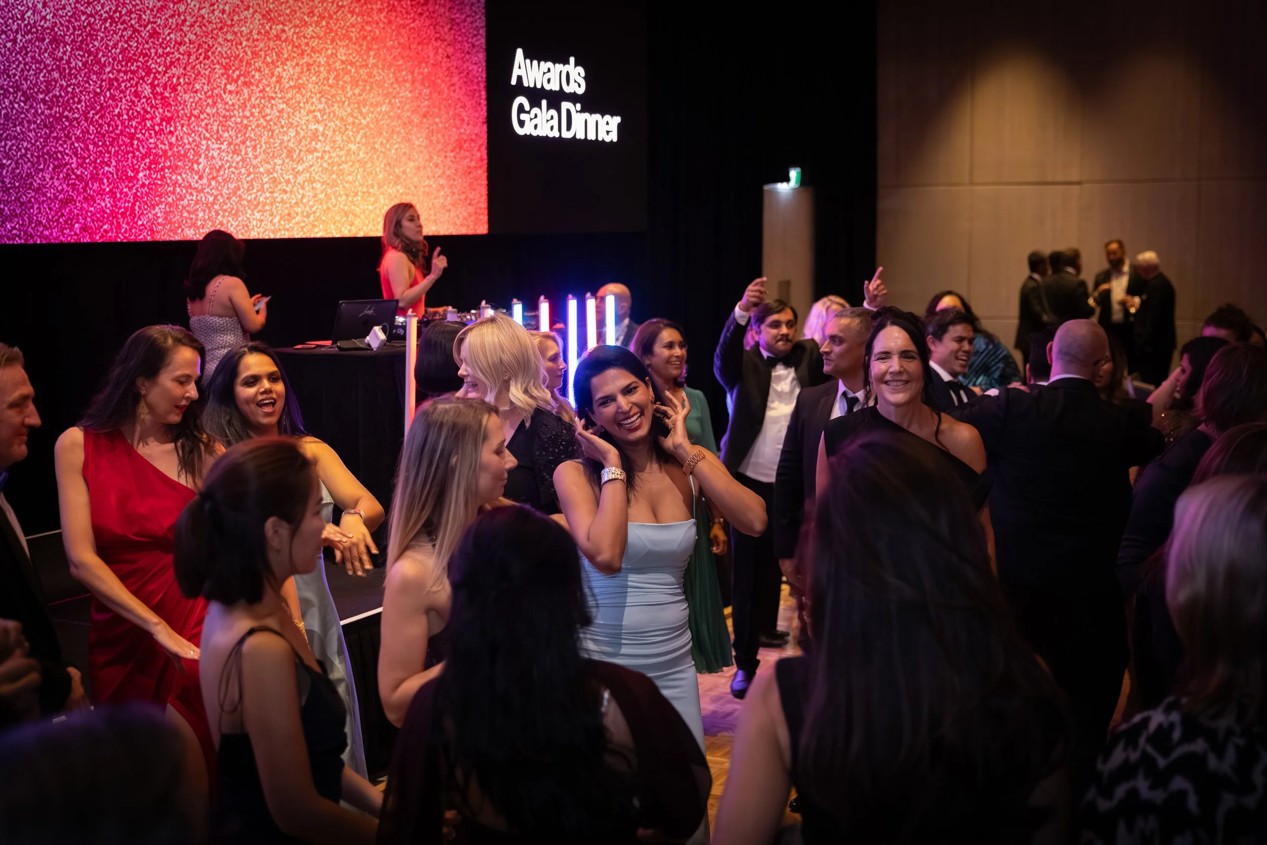 People dancing and enjoying themselves at an awards gala dinner event, with a woman speaking at a podium and a large screen displaying 'Awards Gala Dinner' in the background.