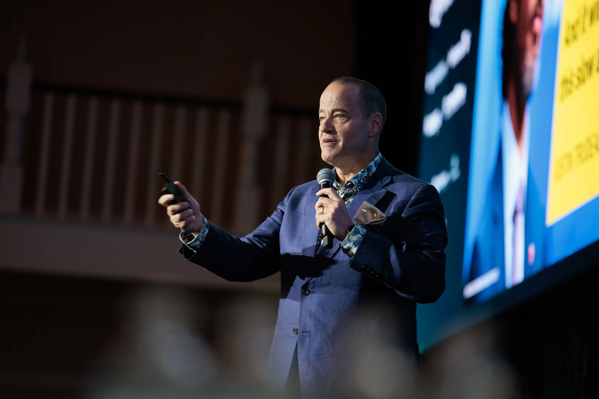 A man in a blue suit holding a microphone and a remote control, speaking at an event with a large screen displaying a colorful presentation behind him.