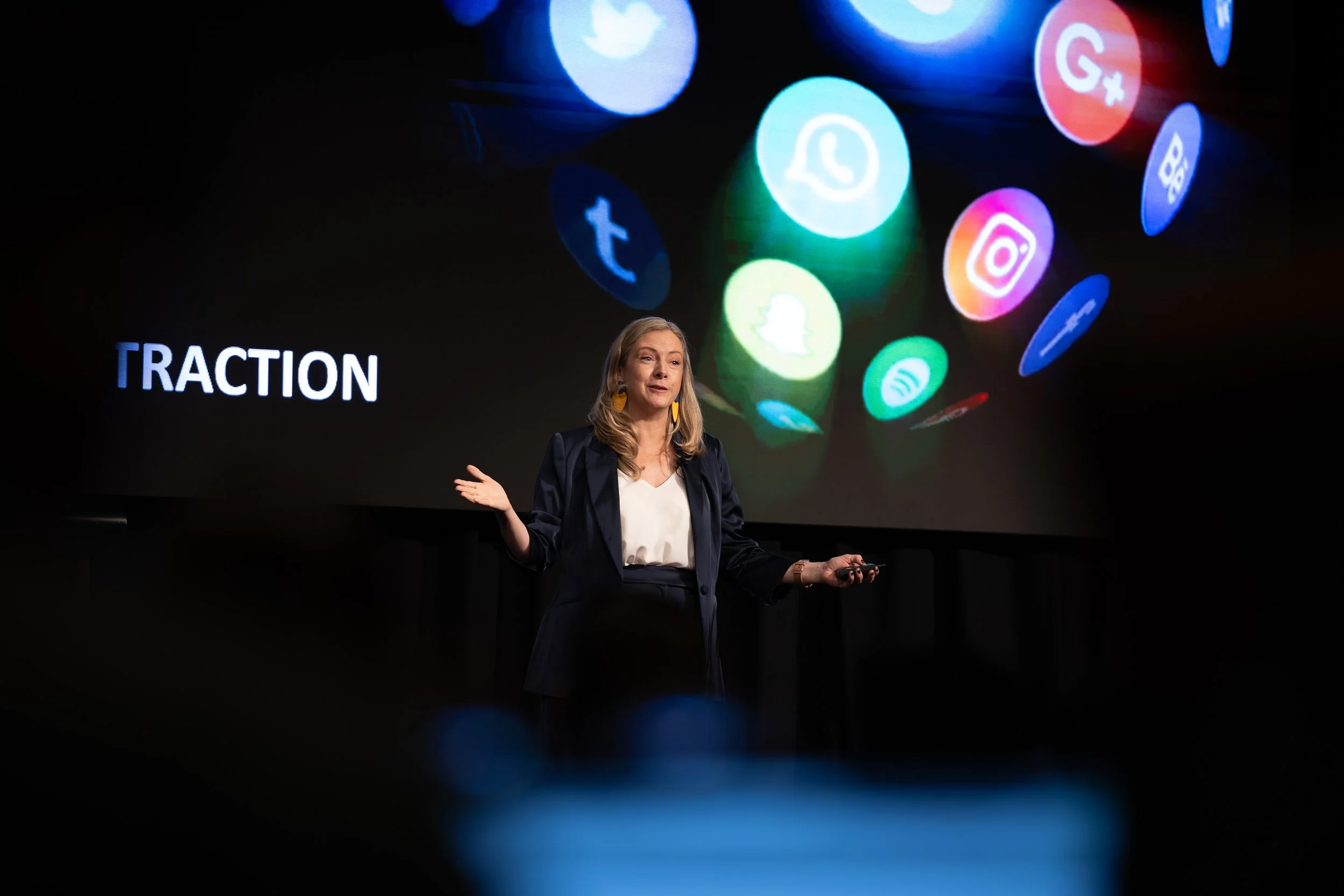 Woman giving a presentation on social media platforms, with a large screen behind her displaying various social media icons and the word 'TRACTION'.