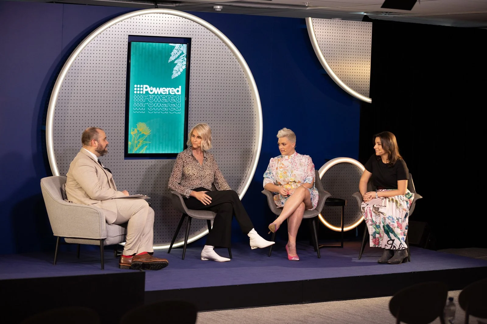 Four people sitting on stage in a discussion panel with a digital screen behind them displaying the word 'Powered' with greenery graphics.