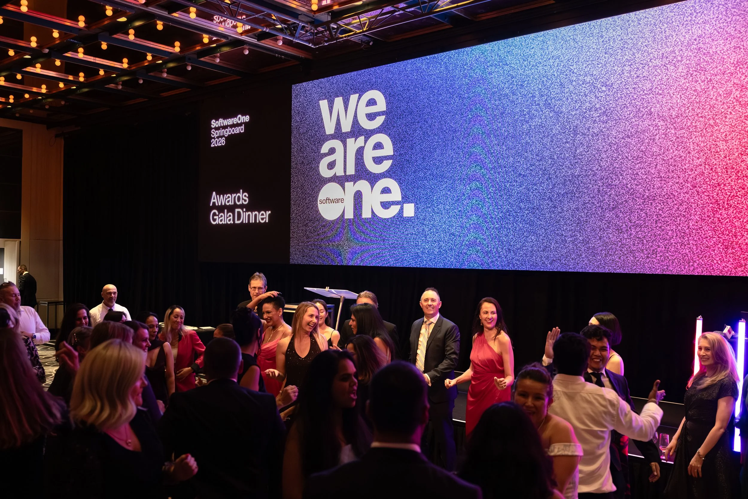 People in formal attire attending an awards ceremony with a large screen displaying the text 'we are one.' and 'SoftwareOne Springboard 2026' in the background.