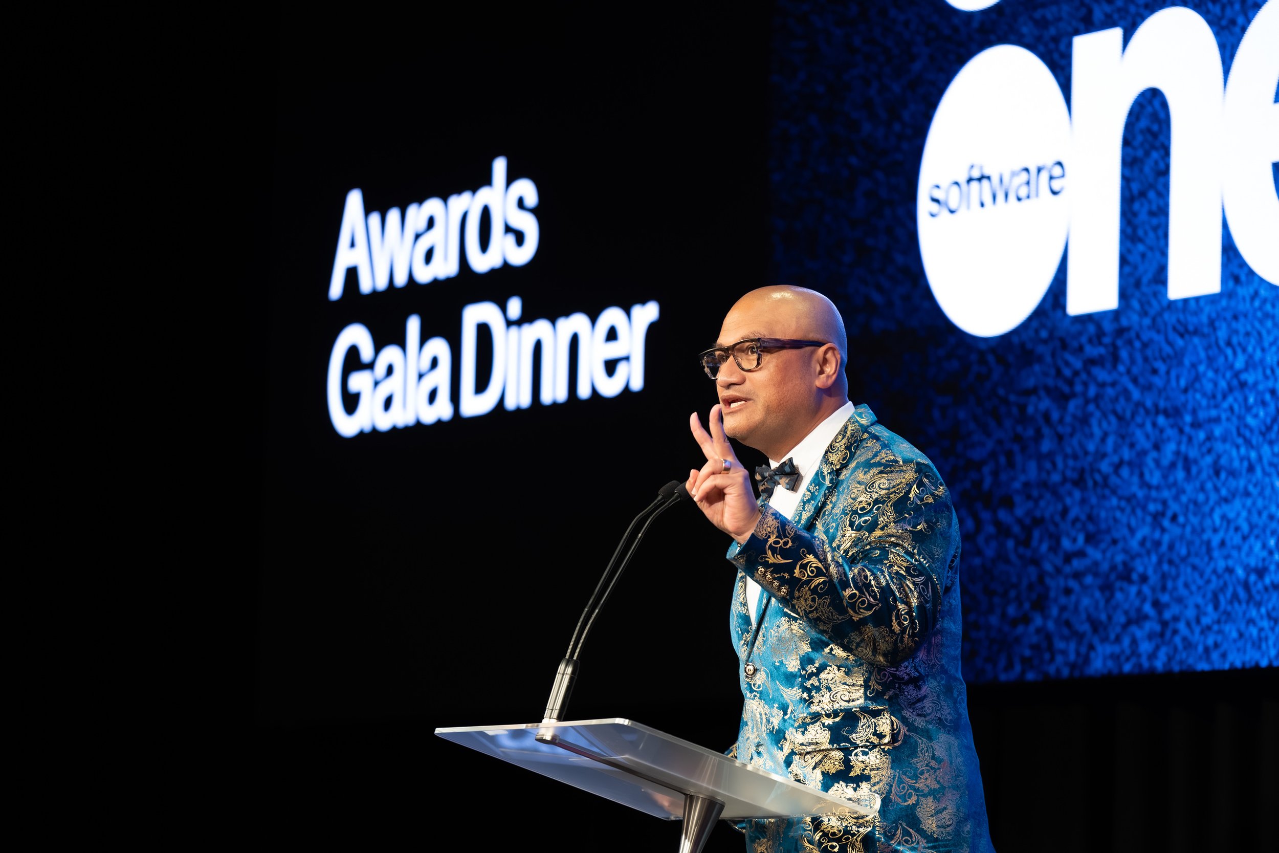 A man in a blue and gold patterned suit speaking at a podium during an awards dinner event, with a large screen behind him displaying the words 'Awards Gala Dinner' and a logo.