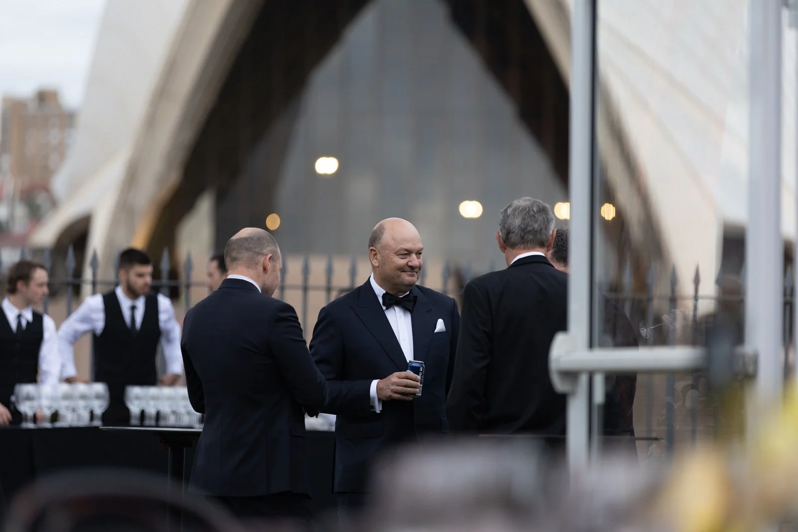 A group of men in formal tuxedos socializing outdoors near the Sydney Opera House, with waitstaff in the background.