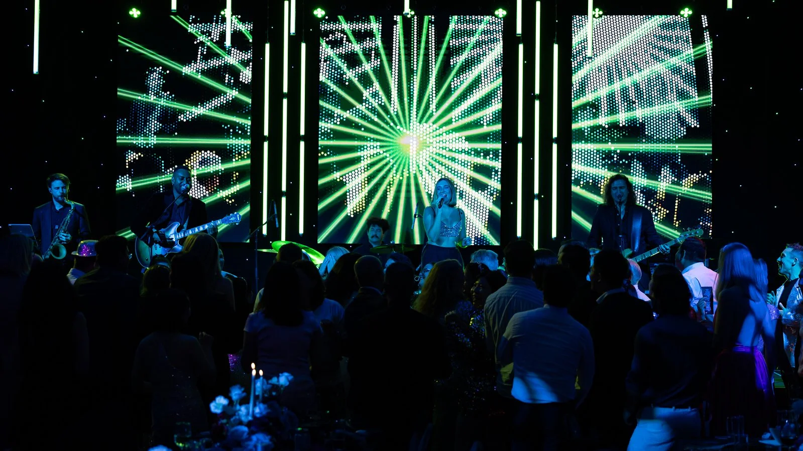 A band performing on stage with a digital LED backdrop displaying green and white lights and abstract patterns. The audience is watching the performance.