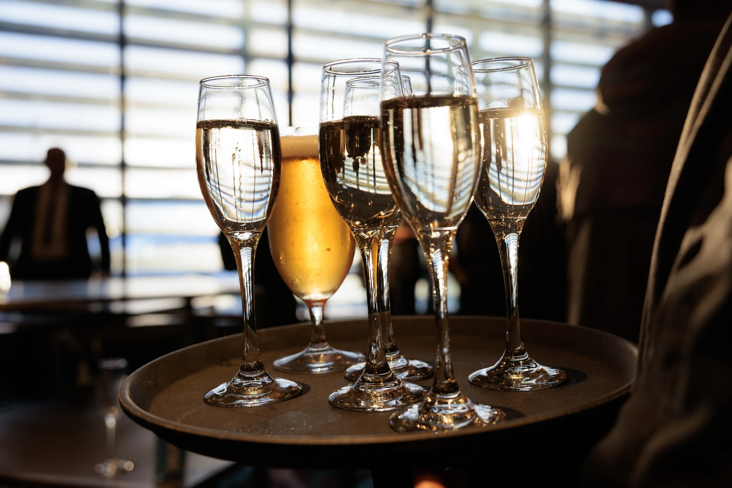 A tray of champagne flutes filled with bubbly on a table, with sunlight reflecting off the glasses in an airport or modern building setting.