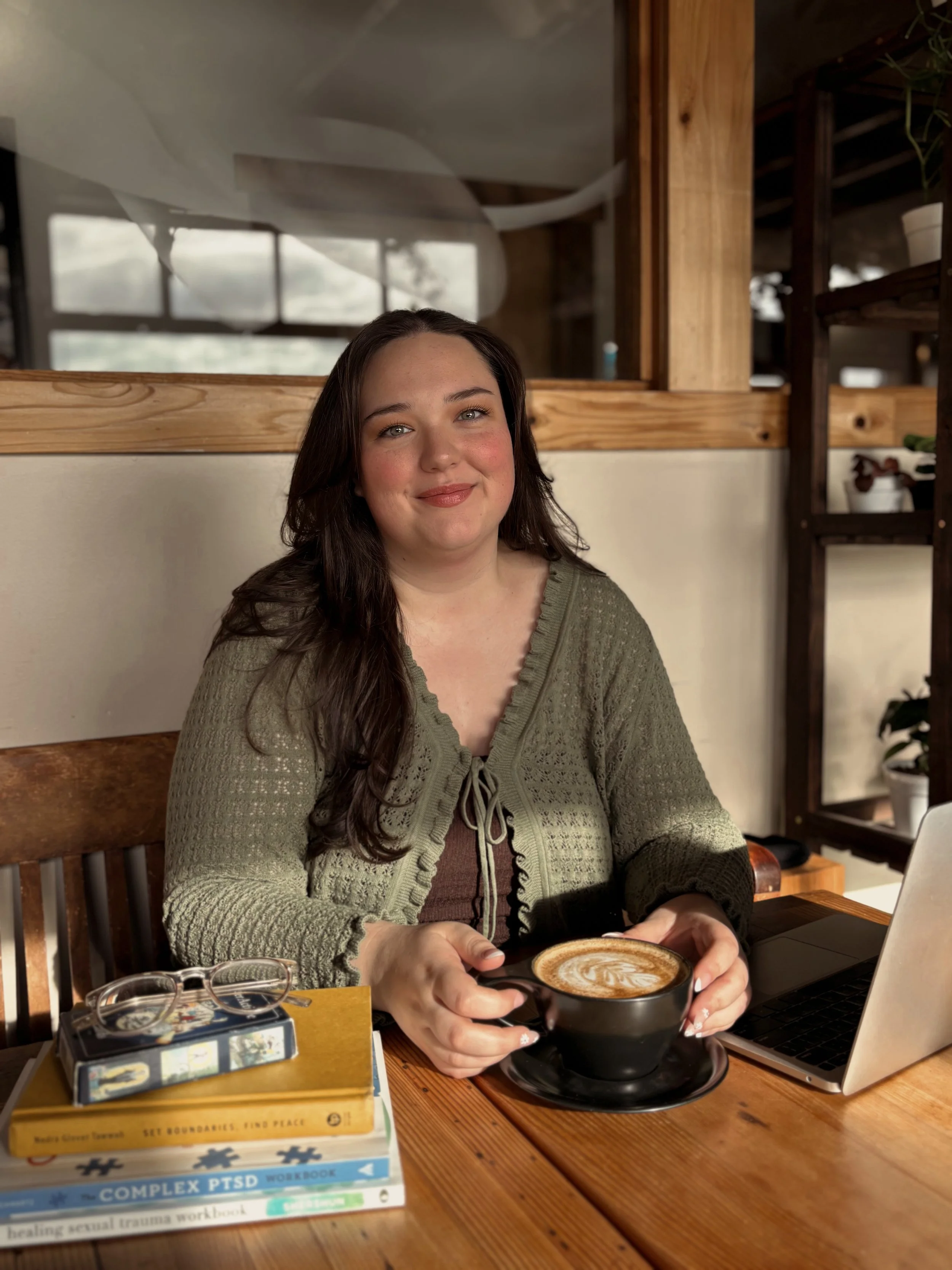 Naomi Stephens, telehealth relational trauma therapist in Oregon and Mississippi, sitting at a table with a stack of books, hot latte, and an open laptop in front of her.