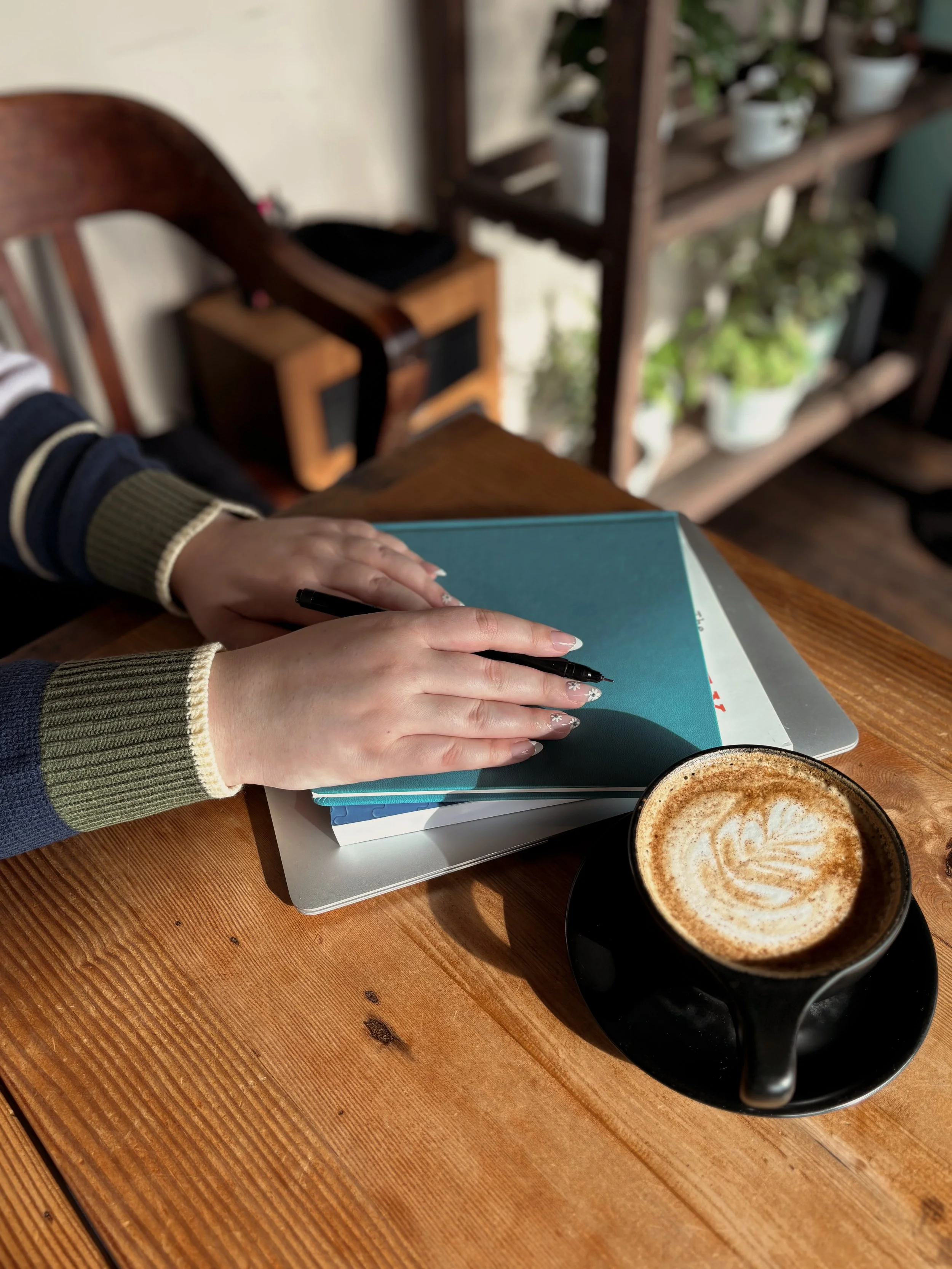 Hands holding a pen while resting on a small stack of books. A hot coffee in a black cup is sitting near. Everything is resting on a wooden table.