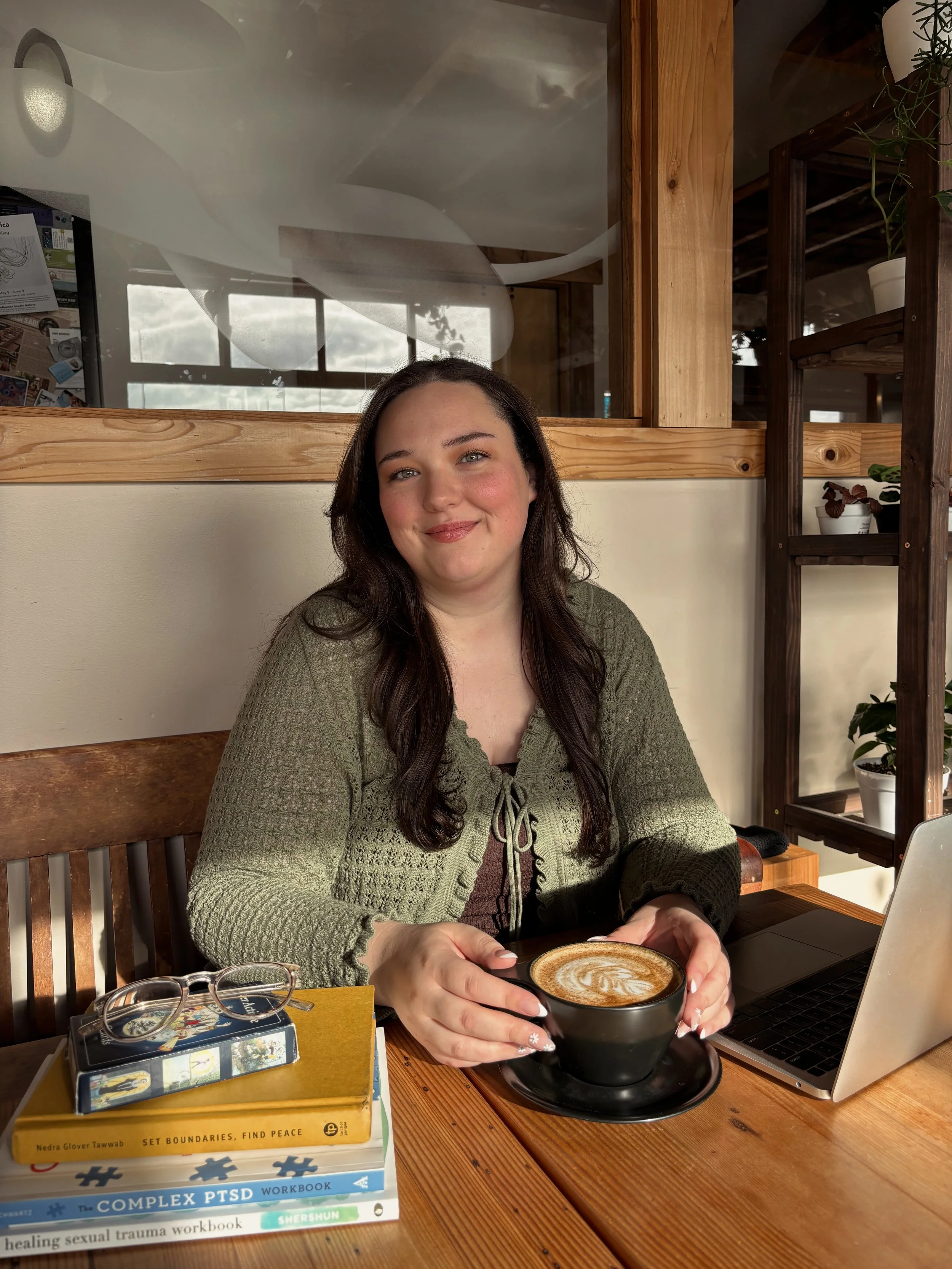 Naomi Stephens sitting at cafe table with books, laptop, and a cup of coffee. She is smiling softly at the camera.