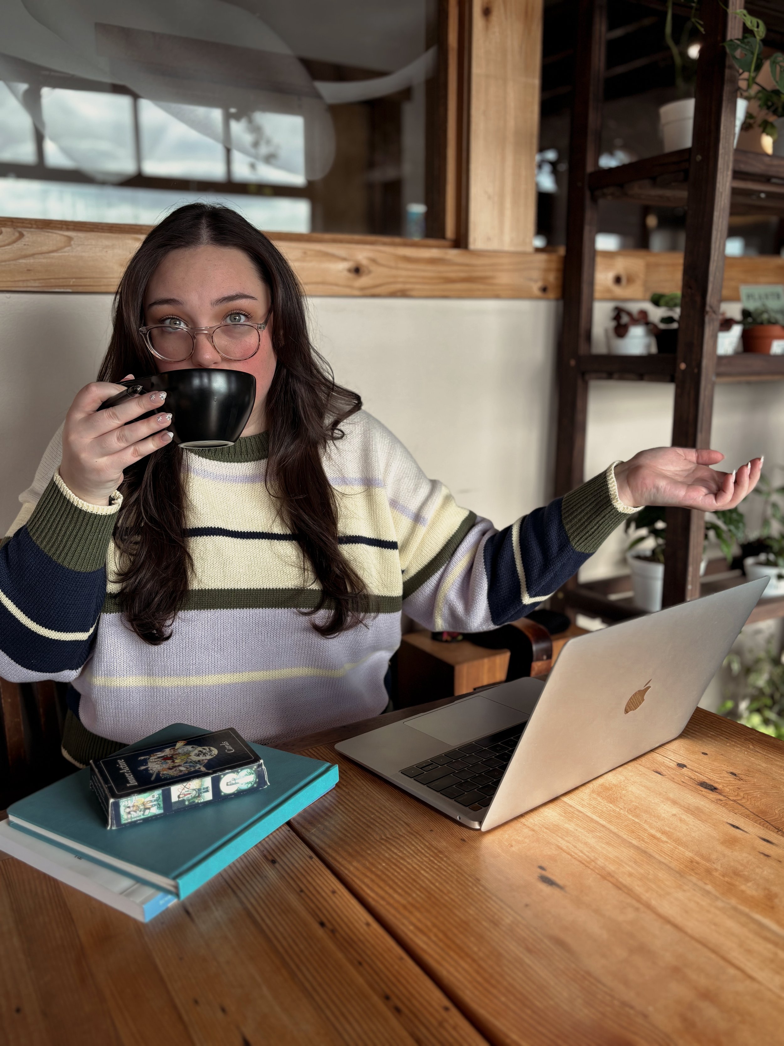 Naomi Stephens, virtual therapist in Oregon, sitting at cafe table with open laptop and small stack of books in front of her. She is sipping from a black coffee cup.