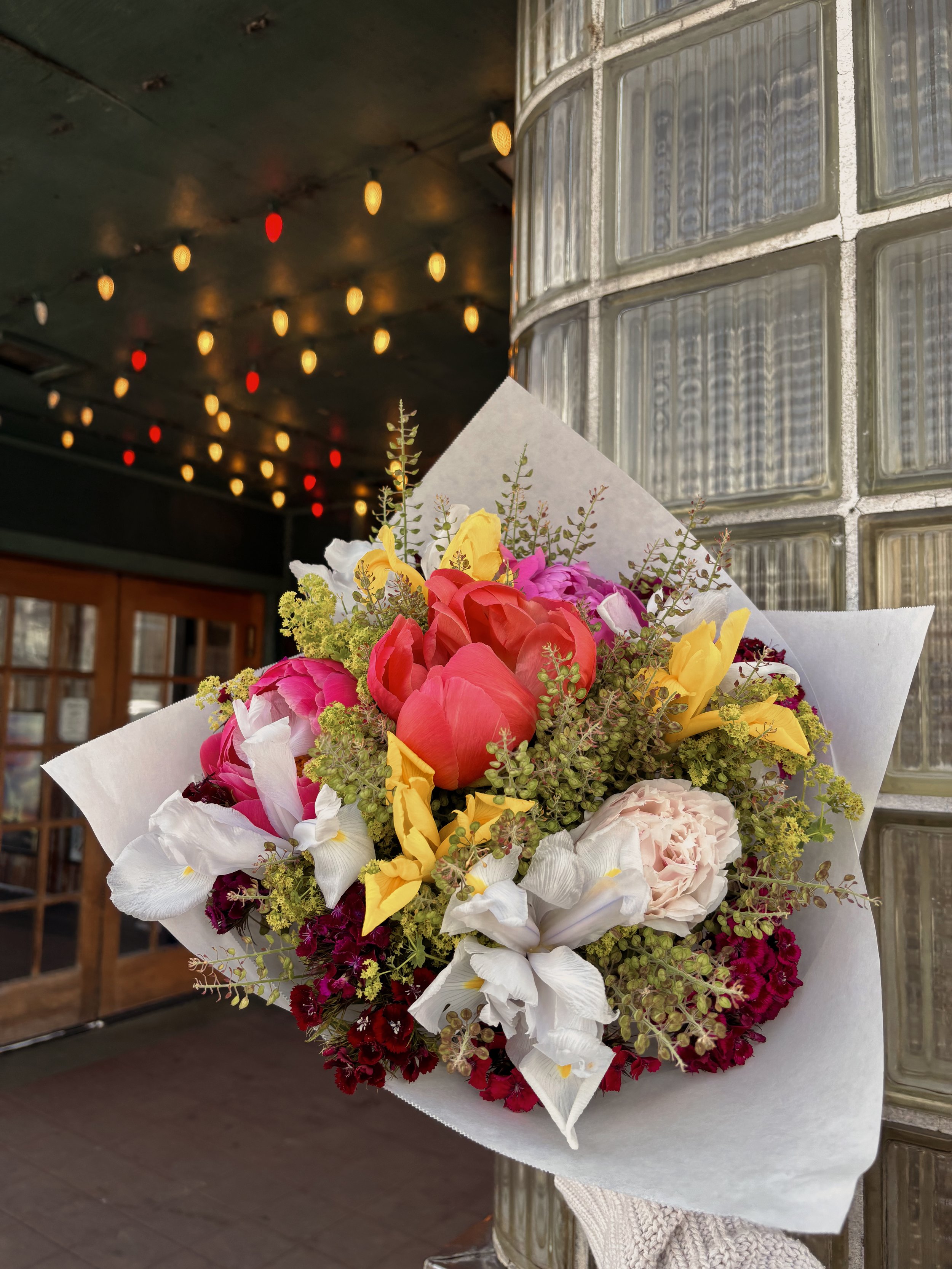 Bouquet of flowers which are colored yellow, pink, white, green, and burgundy. Posed in front of an old theater with warm yellow and red string lights.
