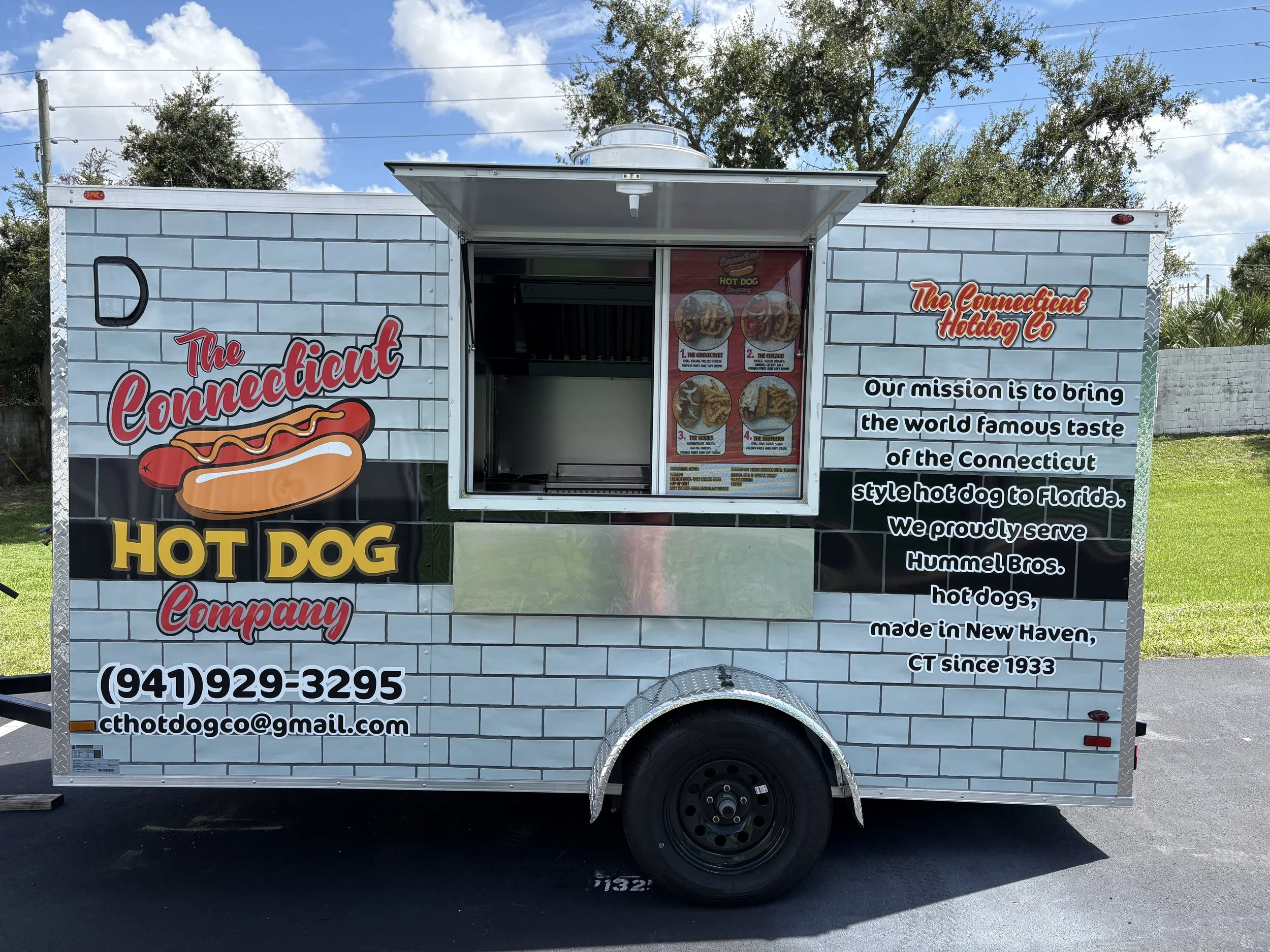 A mobile food trailer decorated with a brick wall pattern, advertising hot dogs. The trailer has a sign for The Connecticut Hotdog Co, with a logo of a hot dog. The side of the trailer displays the company's mission statement and contact information, including a phone number and email. The trailer is parked outdoors under a partly cloudy sky with trees in the background.