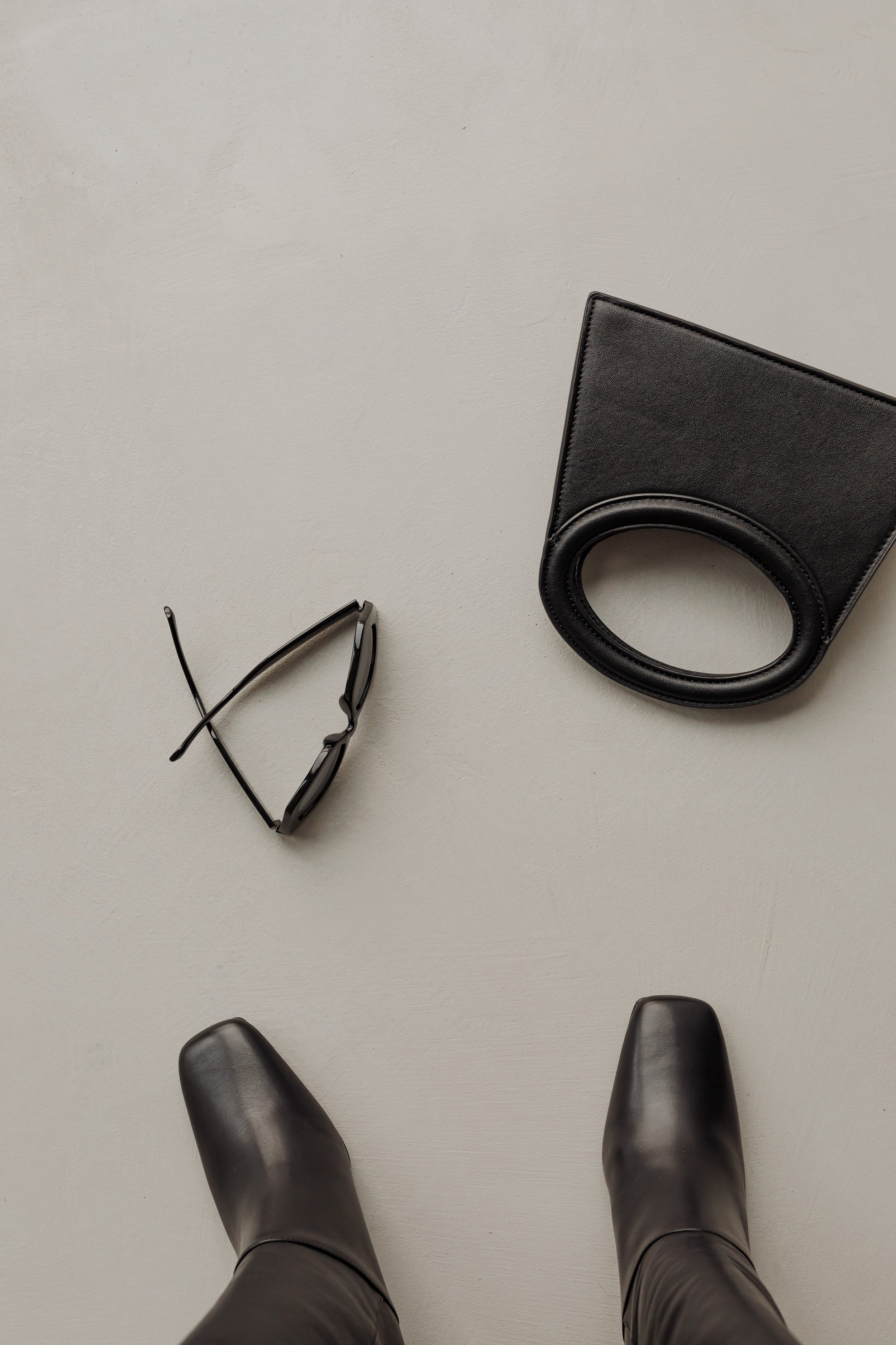 The image shows a pair of black leather boots, black sunglasses, and a black wallet on a white surface.