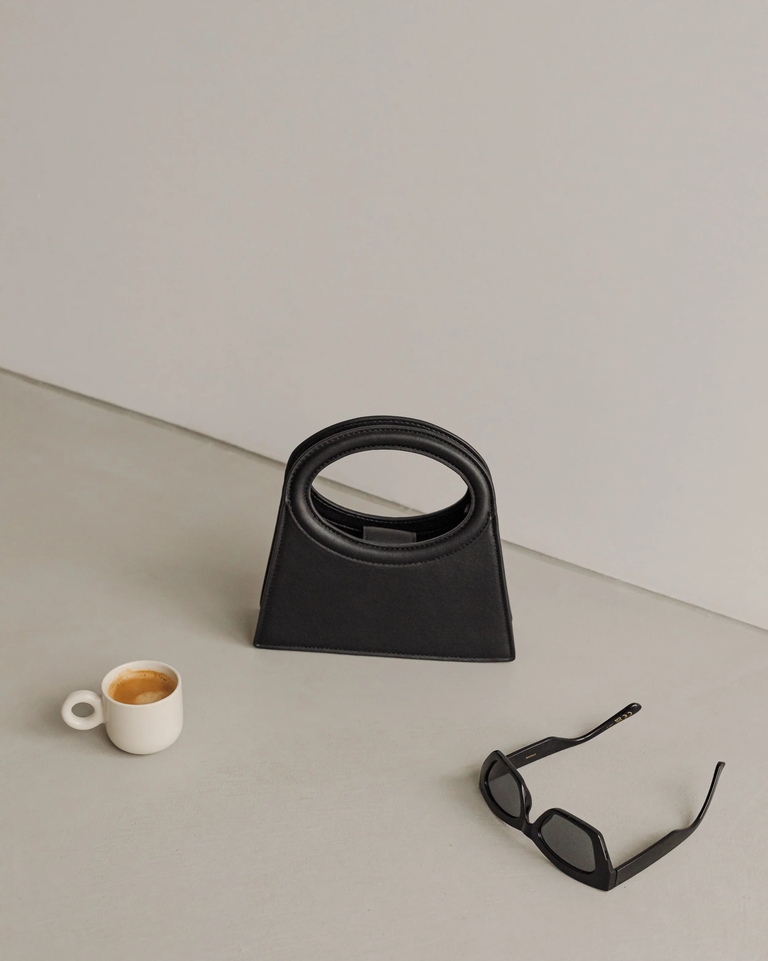 A white coffee mug filled with coffee on a gray table, a pair of black sunglasses and a black purse on the table, with a plain white wall in the background.