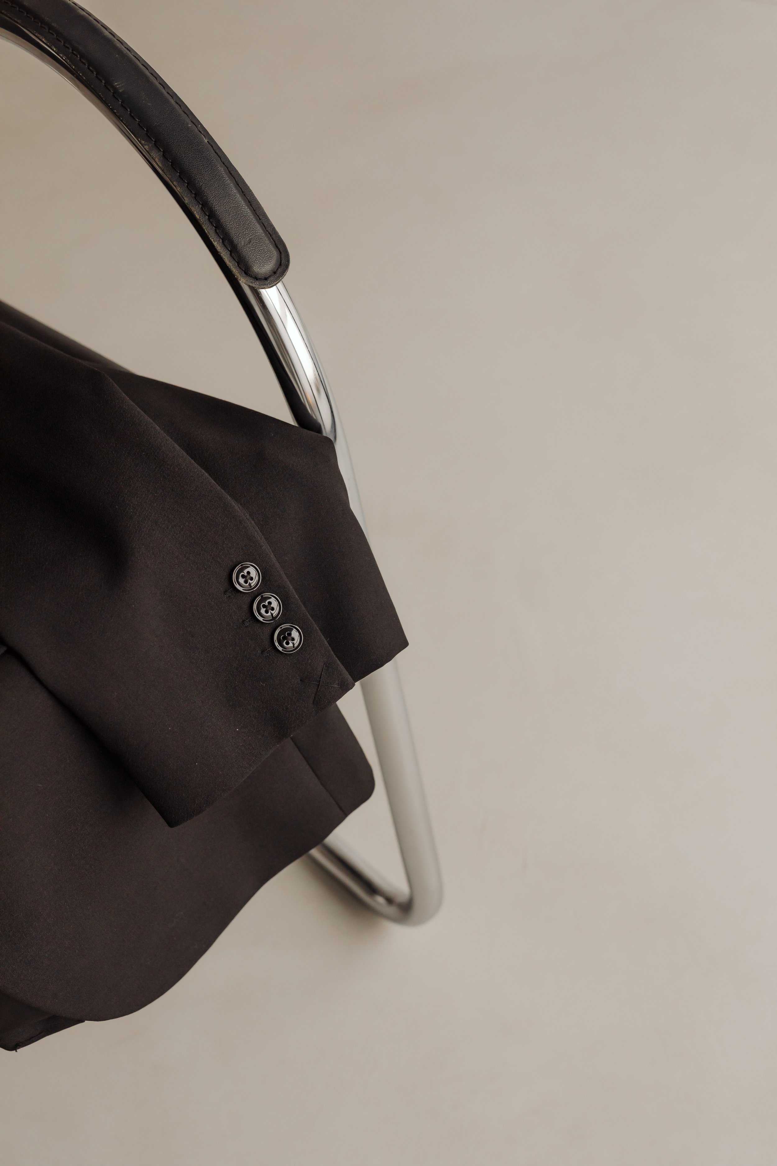 Close-up of a black blazer with four buttons on the sleeve, hanging on a silver chair with a curved backrest, against a plain light-colored wall.