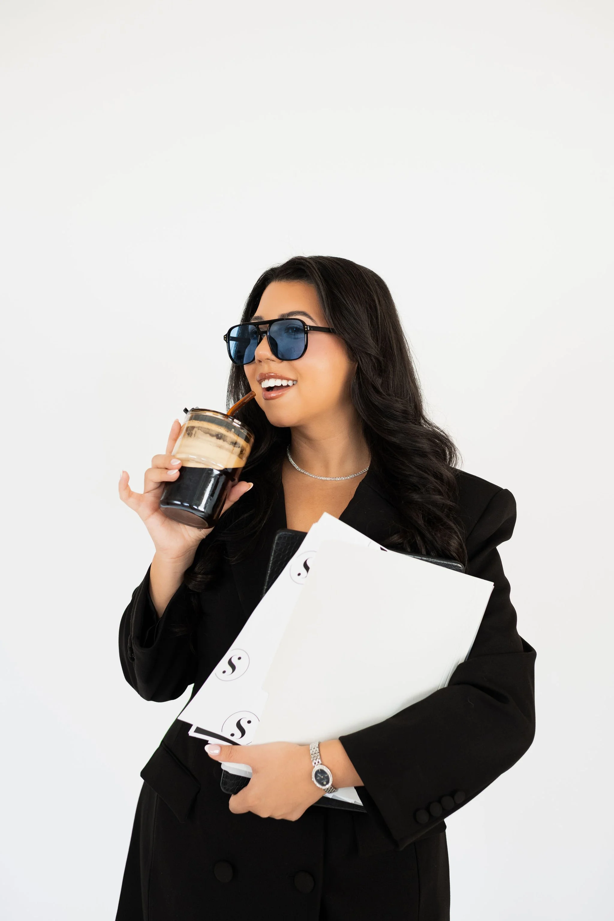 A woman wearing dark sunglasses, a black blazer, holding a coffee drink, and holding a white folder with a 'S' logo, against a plain white background.