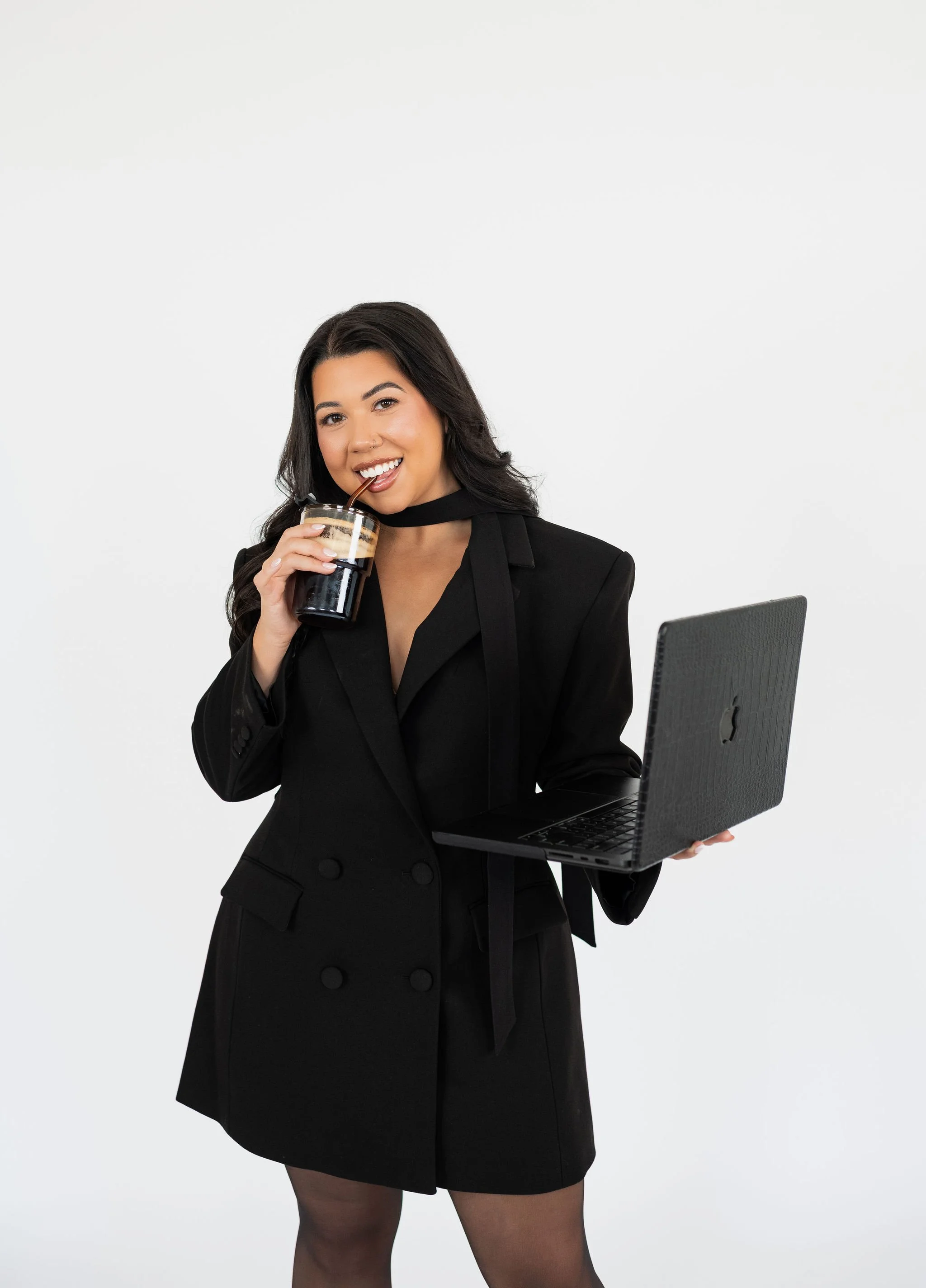 A young woman in a black blazer and skirt, with dark hair and makeup, holding a laptop in one hand and sipping a drink through a straw, standing against a plain white background.