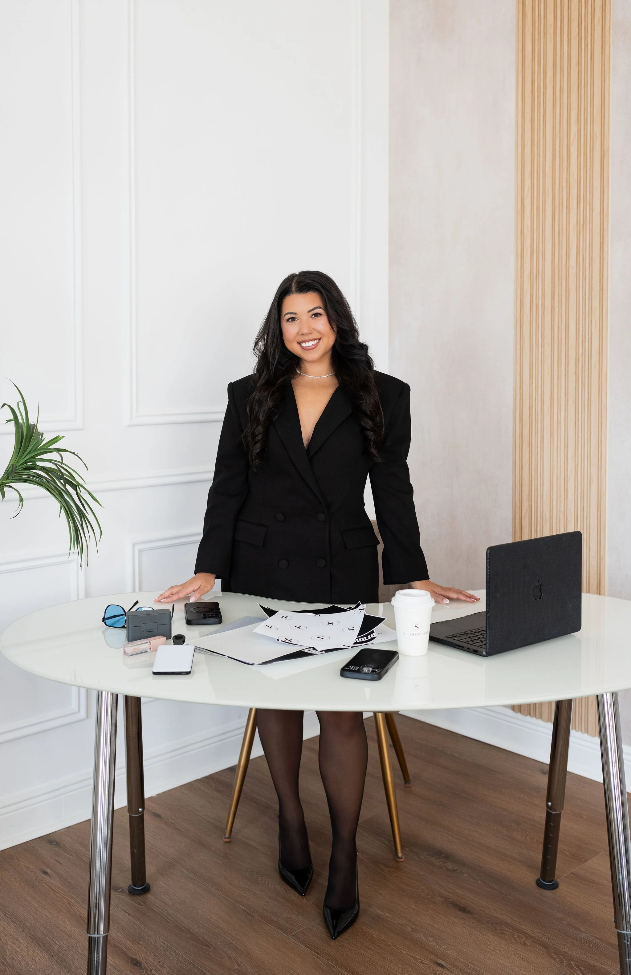A woman with long dark hair in a black blazer, standing behind a white table in a modern office. The table has electronic devices, papers, and a coffee cup. She is smiling and looking at the camera, with the office wall and a plant visible in the background.