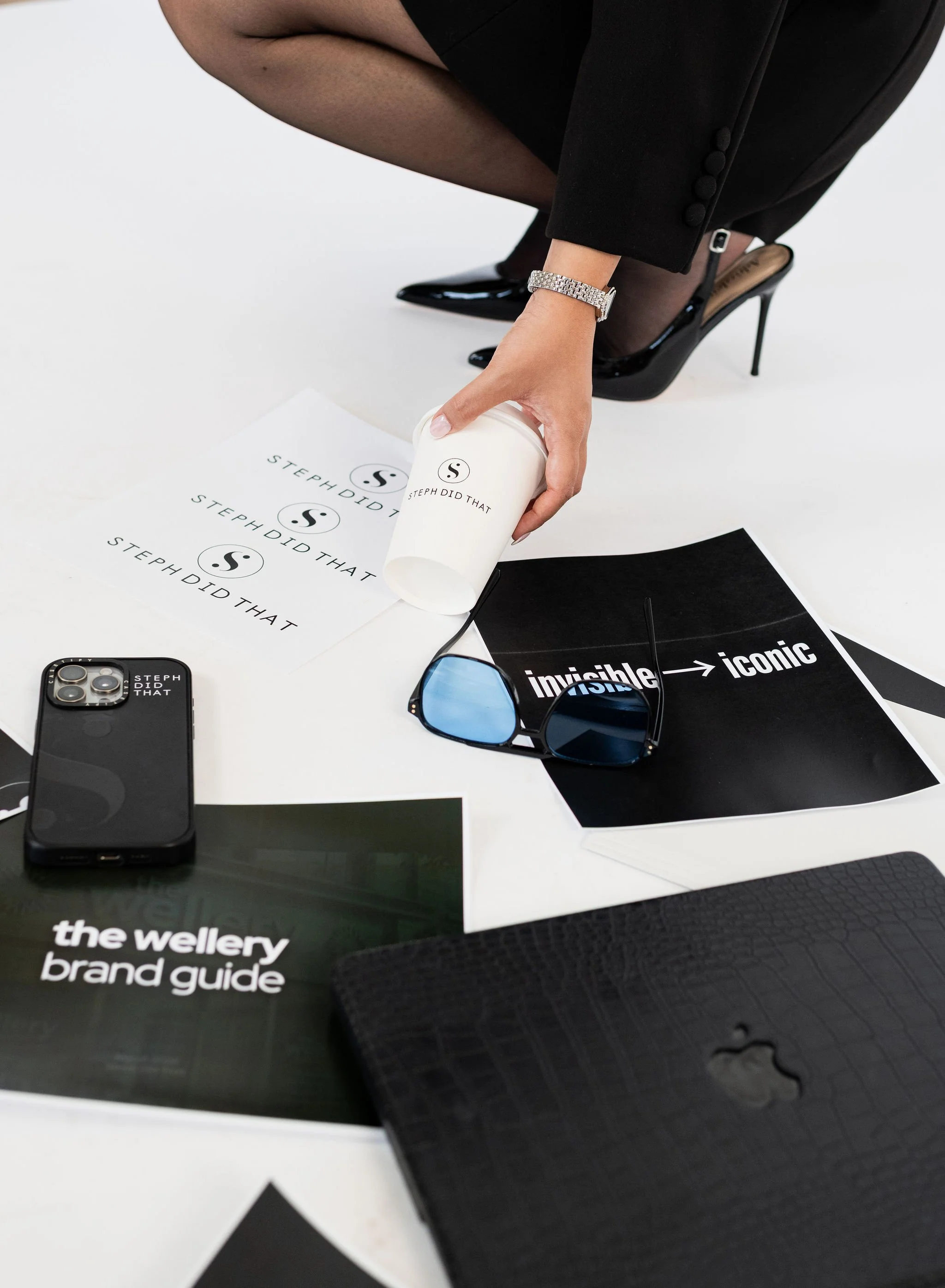 Close-up of a person's hand placing a white paper cup with text 'STEPHDID THAT' on a table with printed materials and a black phone.