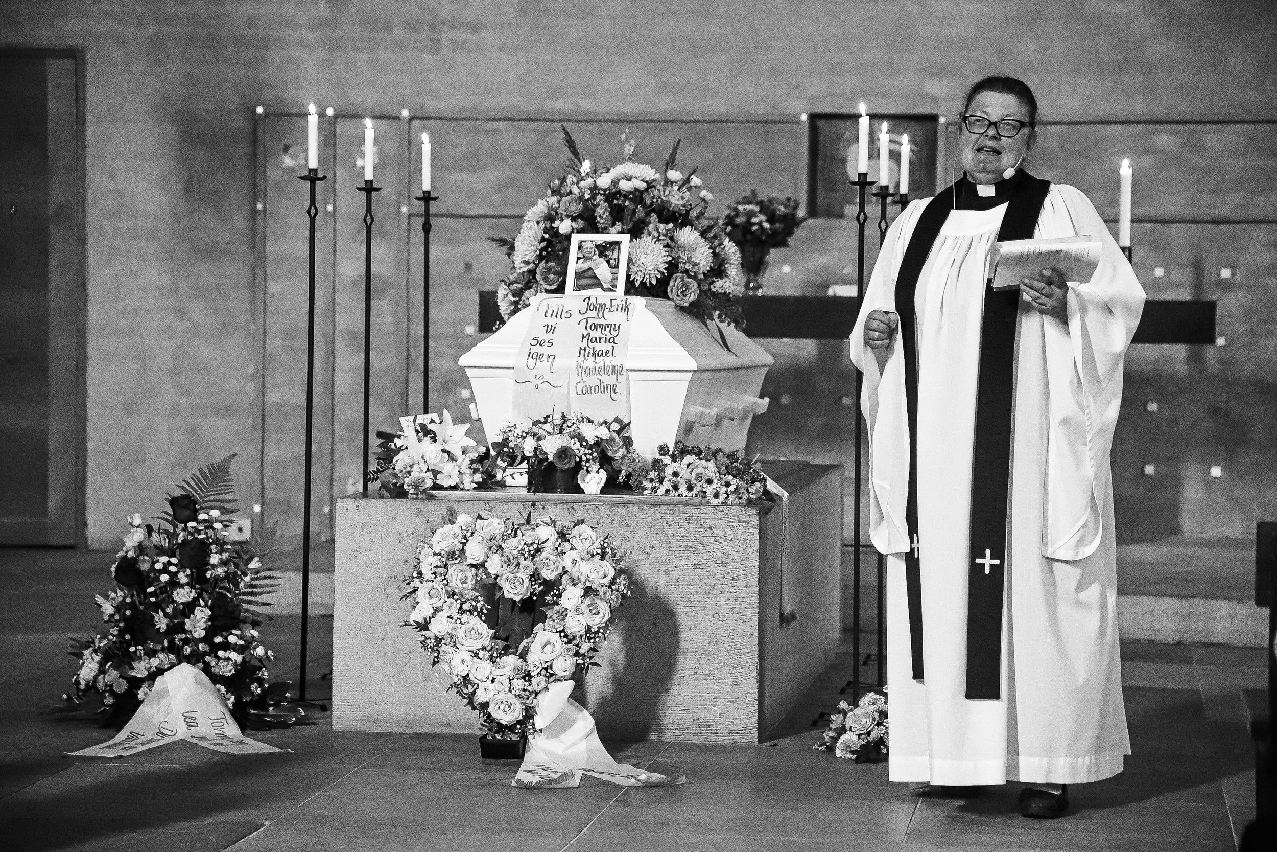 En präst talar informellt i en kyrka, omgiven av blommor och ljus.