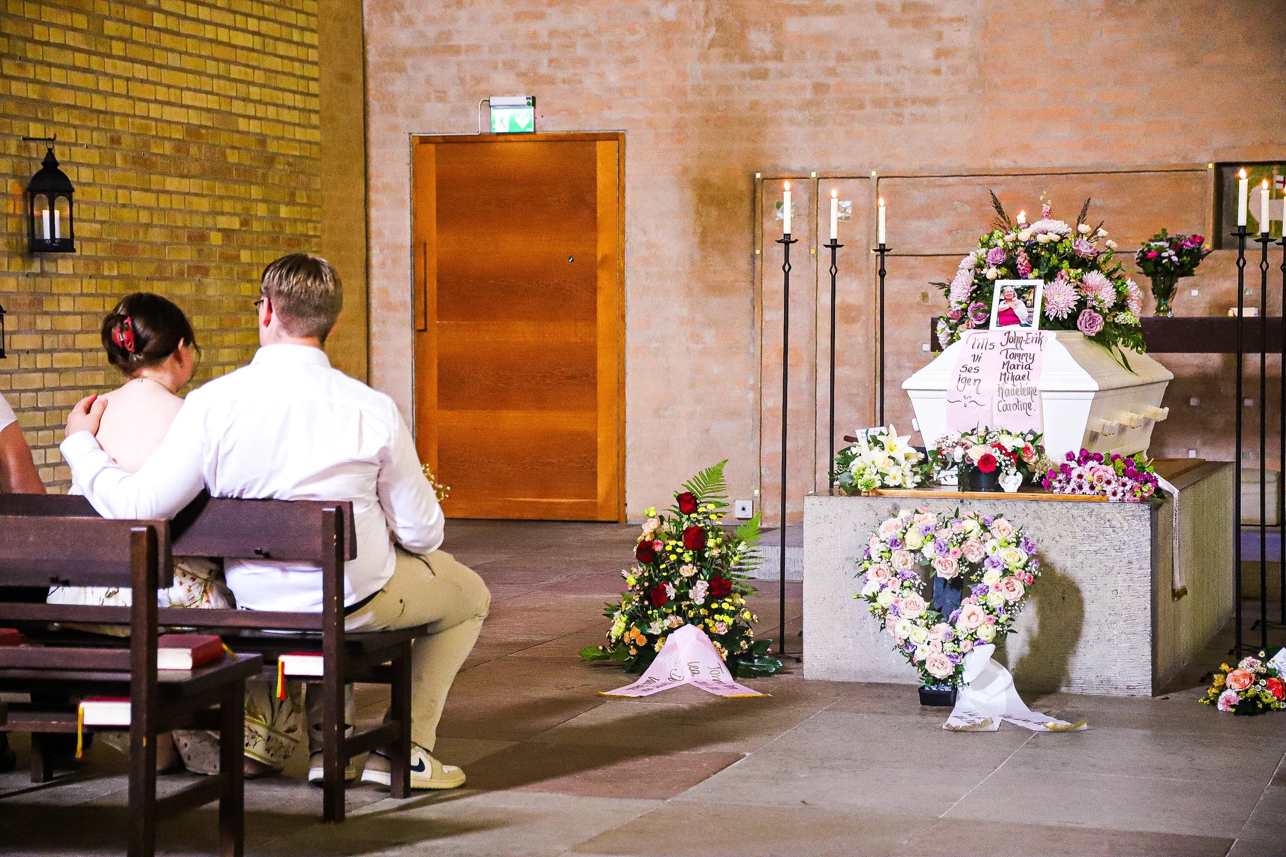 Två personer sitter på bänkar framför en kista dekorerad med många blommor och ett fotografi av en äldre kvinna, i ett kapell för en begravning.