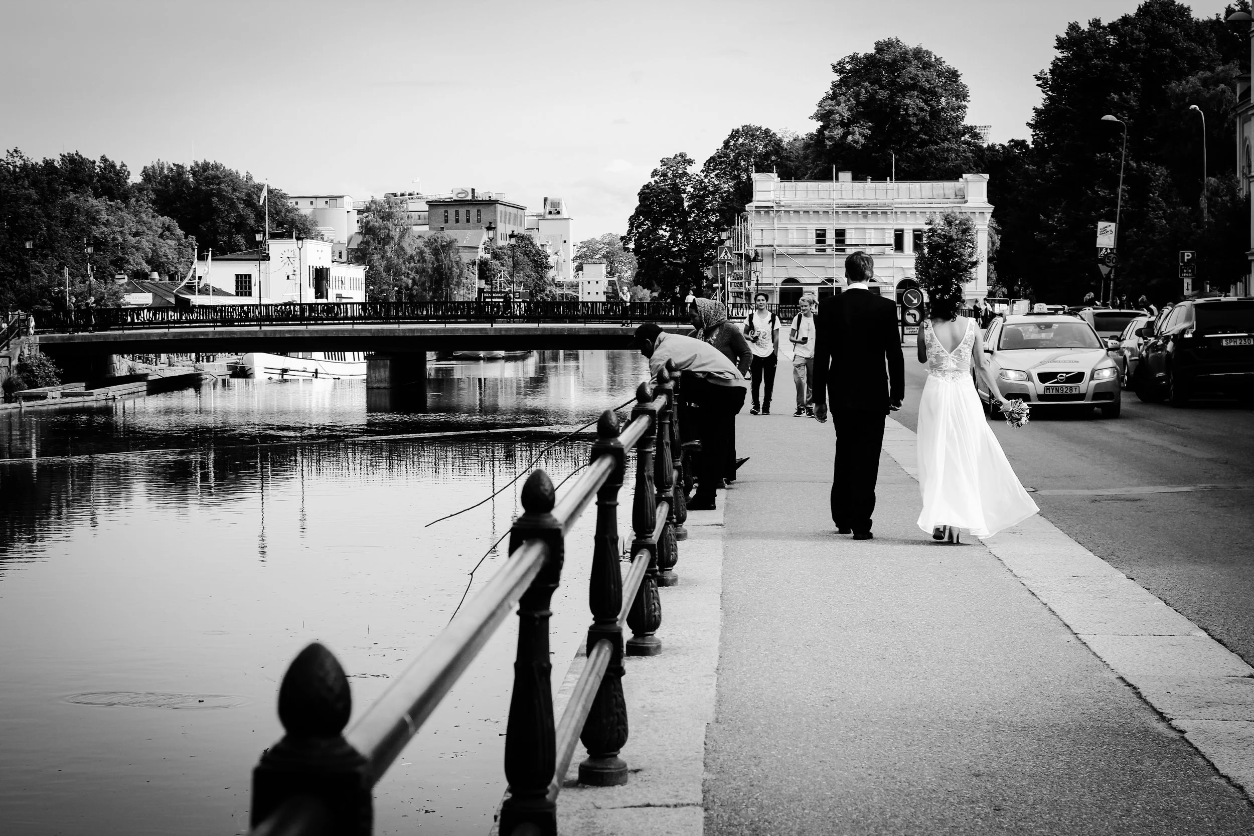 En svartvit bild av en stadsvy med en kanal, en bro och människor på promenadväg. En person i en bröllopsklänning och en man i kostym går tillsammans, medan andra personer står och fiskar vid vattnet. Monuments, byggnader och träd syns i bakgrunden.