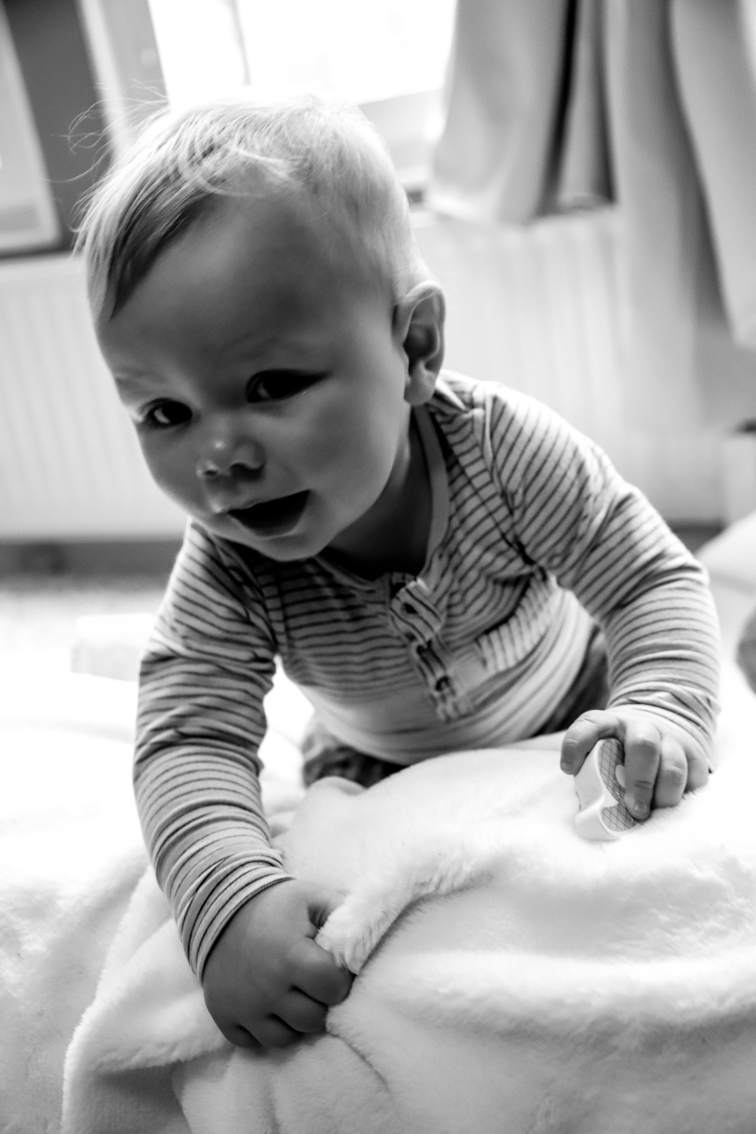 En ung pojke med ljus hår i rand, som klättrar på en mjuk vit yta, troligen ett täcke eller filt, och tittar mot kameran med ett öppet leende.