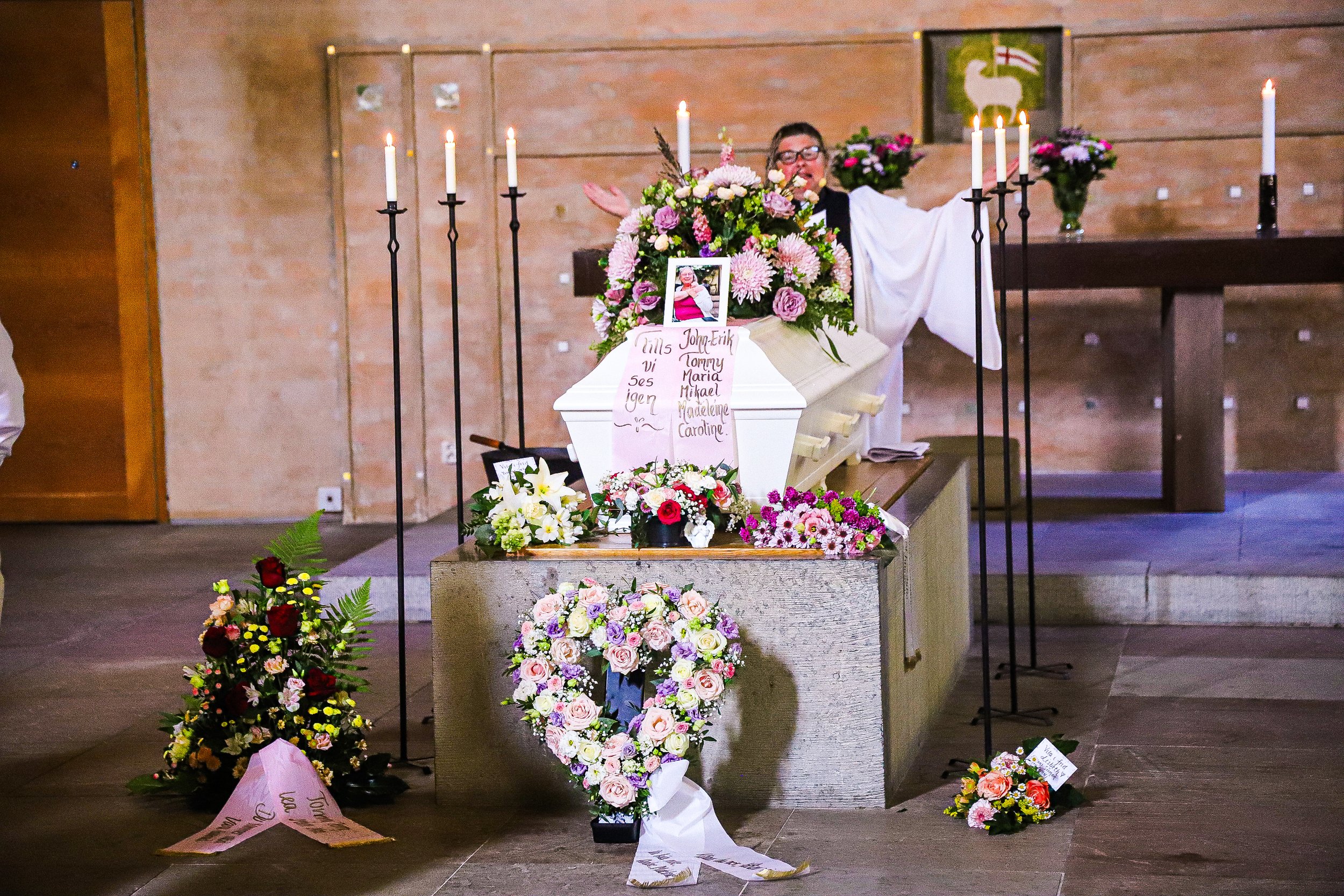 En kista omgiven av många blomsterarrangemang och ljus i en kyrka, tillägnad någon som har gått bort.