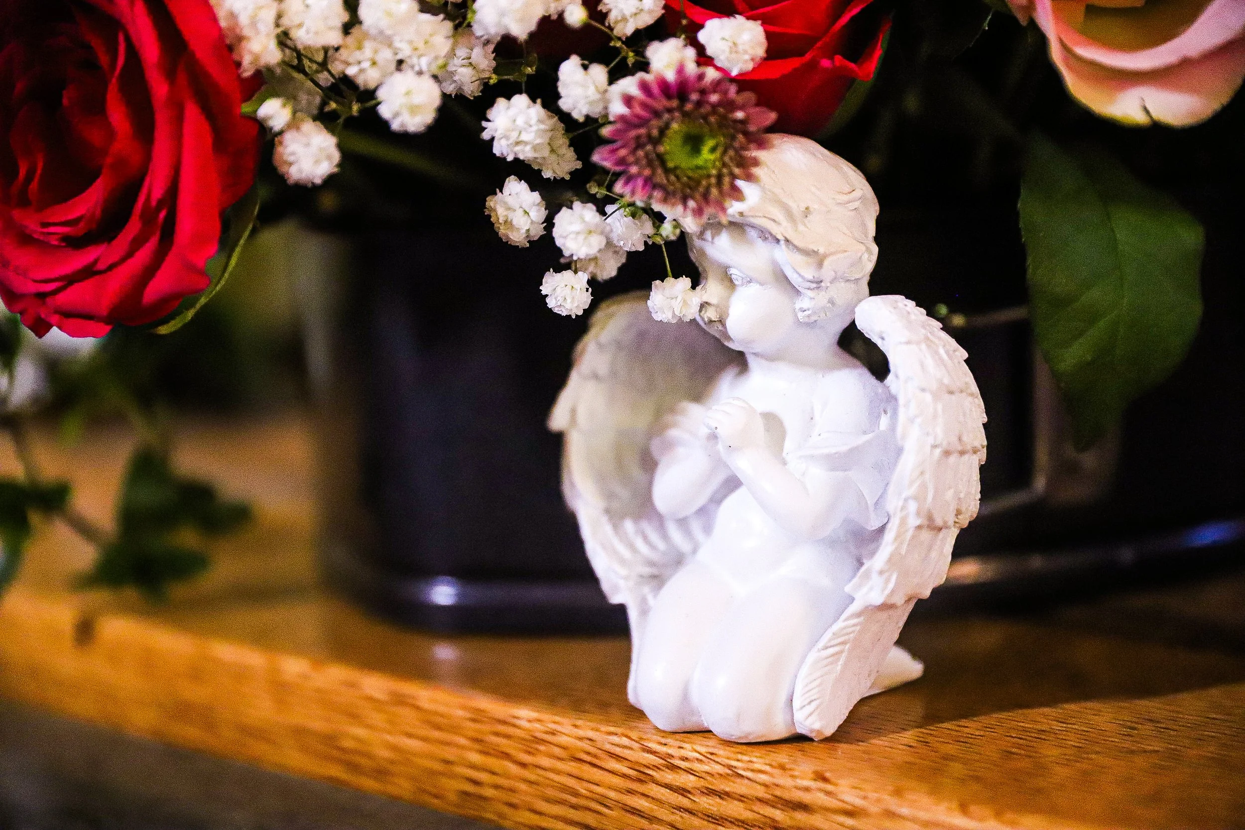 En vit skulptur av en ängel med blommor på huvudet, placerad på ett träbord med blommor i bakgrunden.