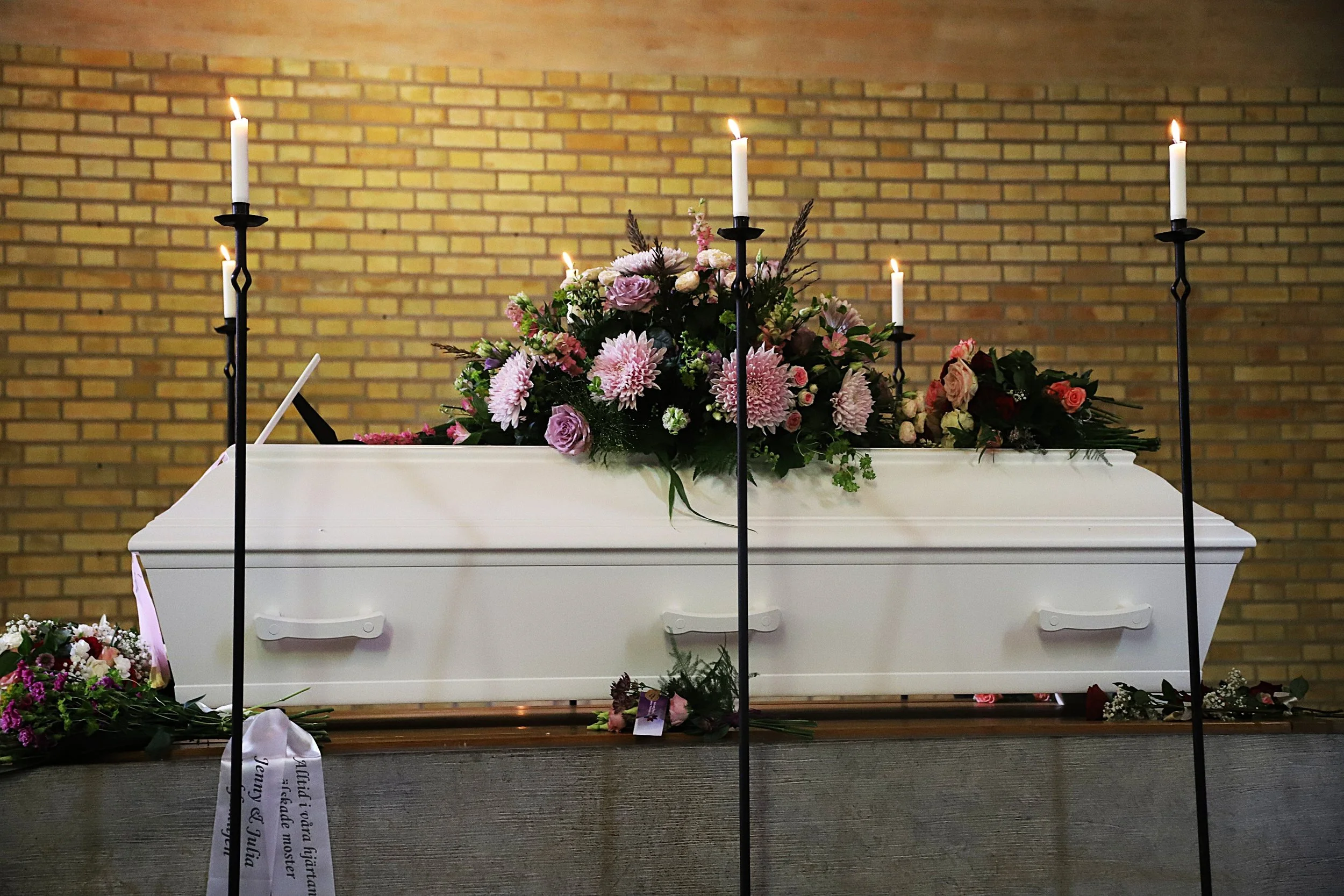 En vit kista med blomsterarrangemang av rosa och vita blommor, omgiven av blomsterarrangemang och ljus i en kyrka eller kapell.