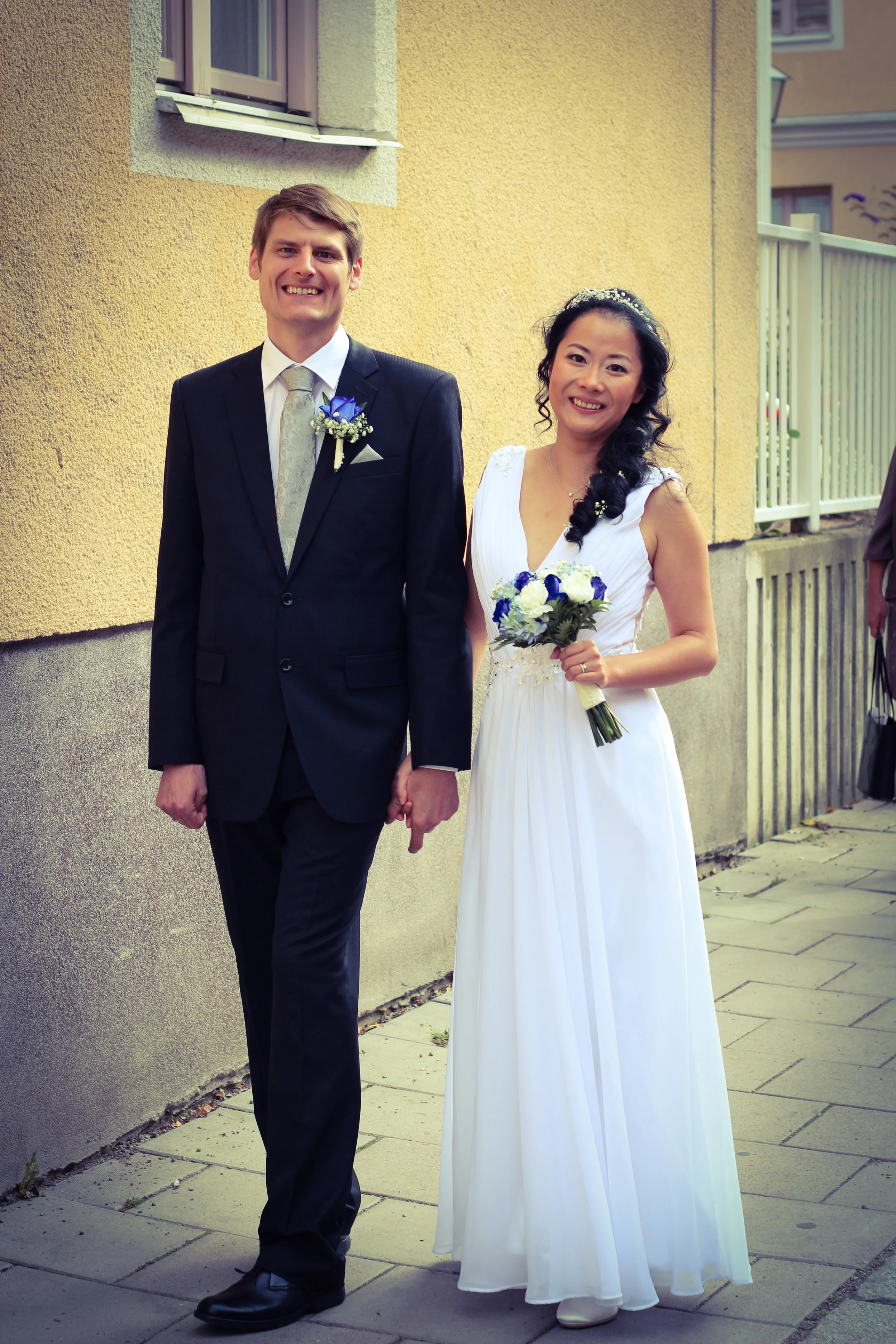 En man och en kvinna i bröllopskläder håller handen och står på trottoaren utomhus, glatt leende, framför en gul vägg och ett fönster med fönsterluckor.